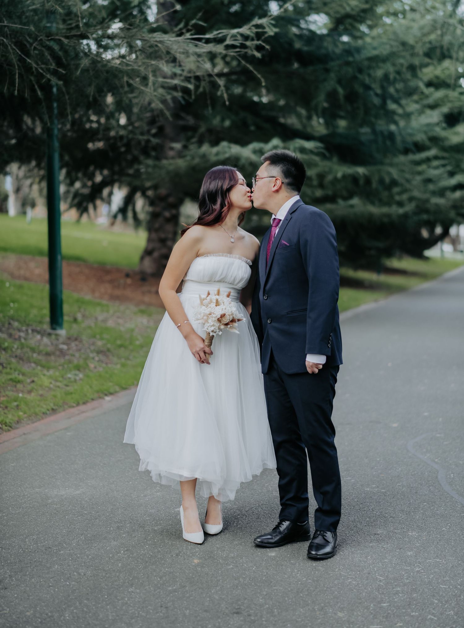 A couple in wedding attire shares a romantic moment on a tree-lined path in the park.