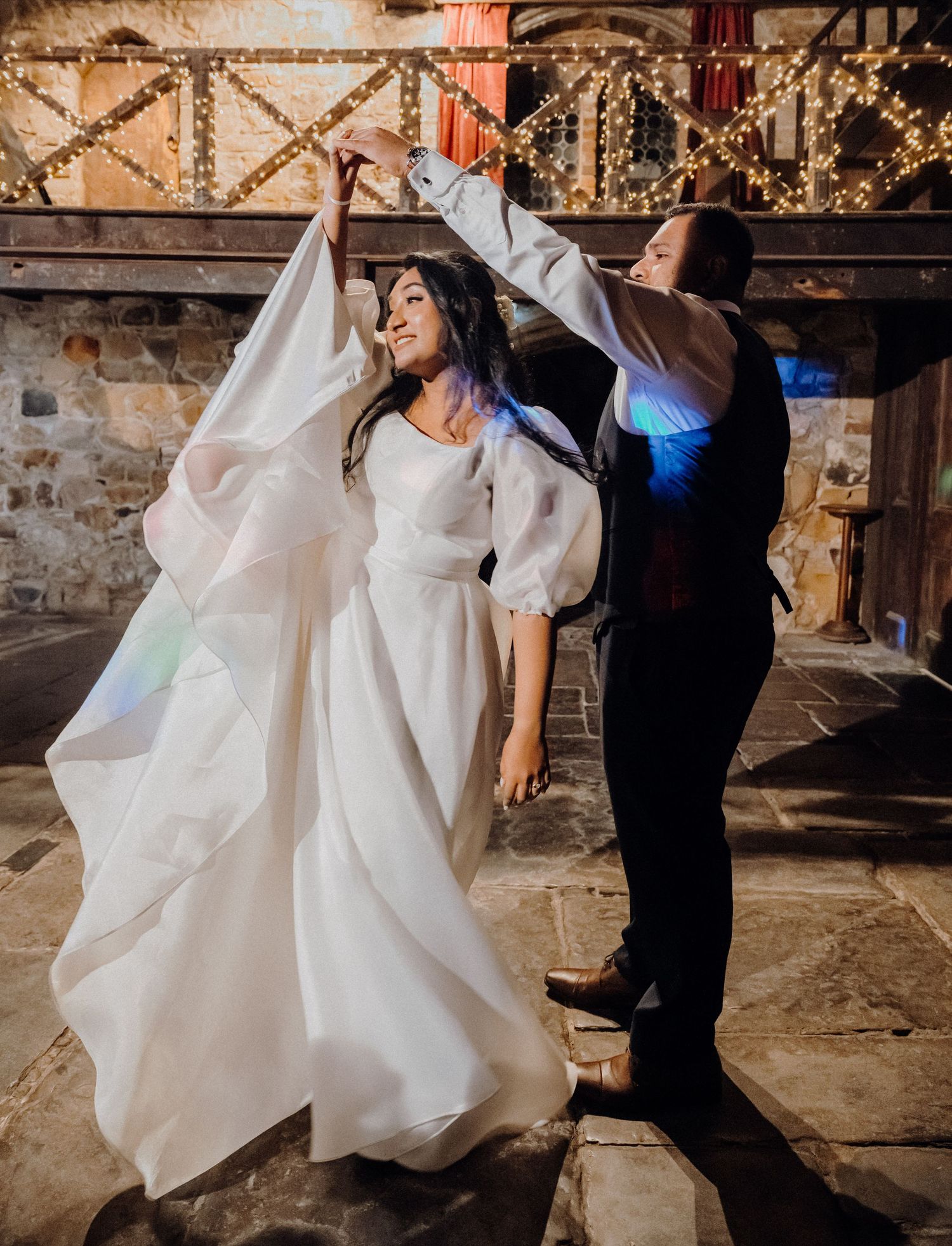 A couple dances together at a rustic wedding reception venue decorated with string lights and stone walls.