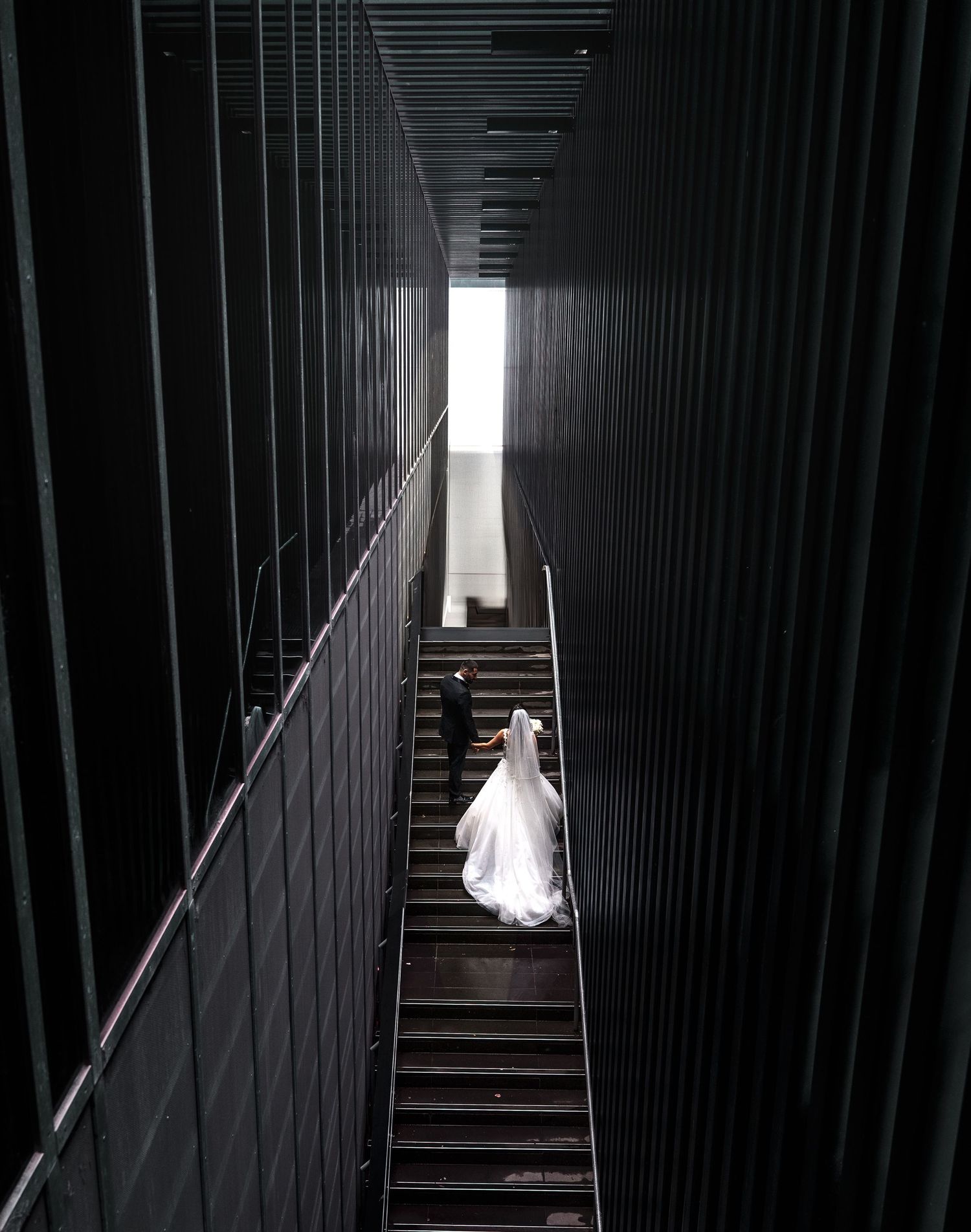 A figure in a flowing white wedding dress descends dark industrial stairs between towering vertical walls leading to light.