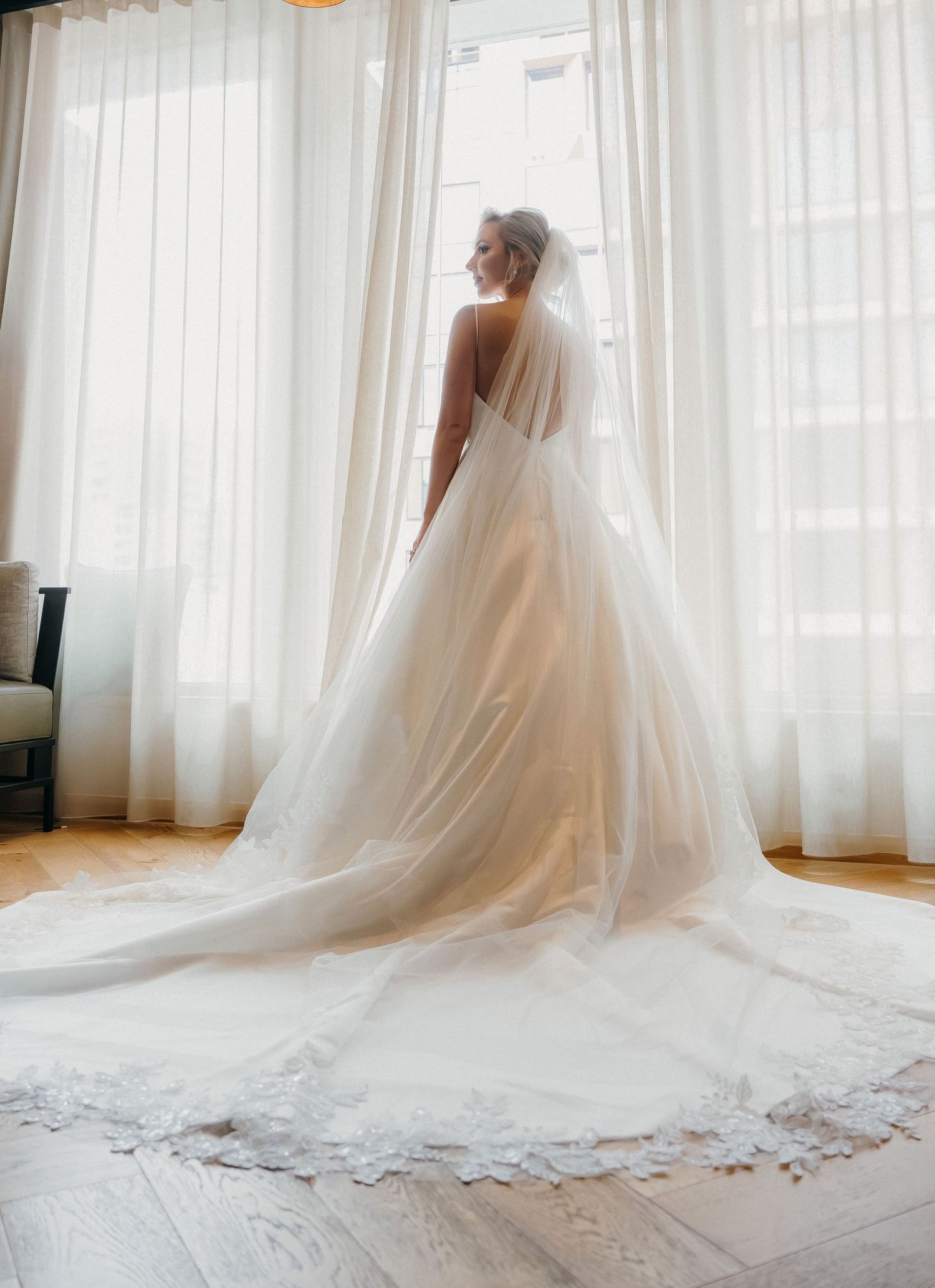 A flowing white wedding gown with cathedral-length veil creates an ethereal silhouette against sheer curtained windows.
