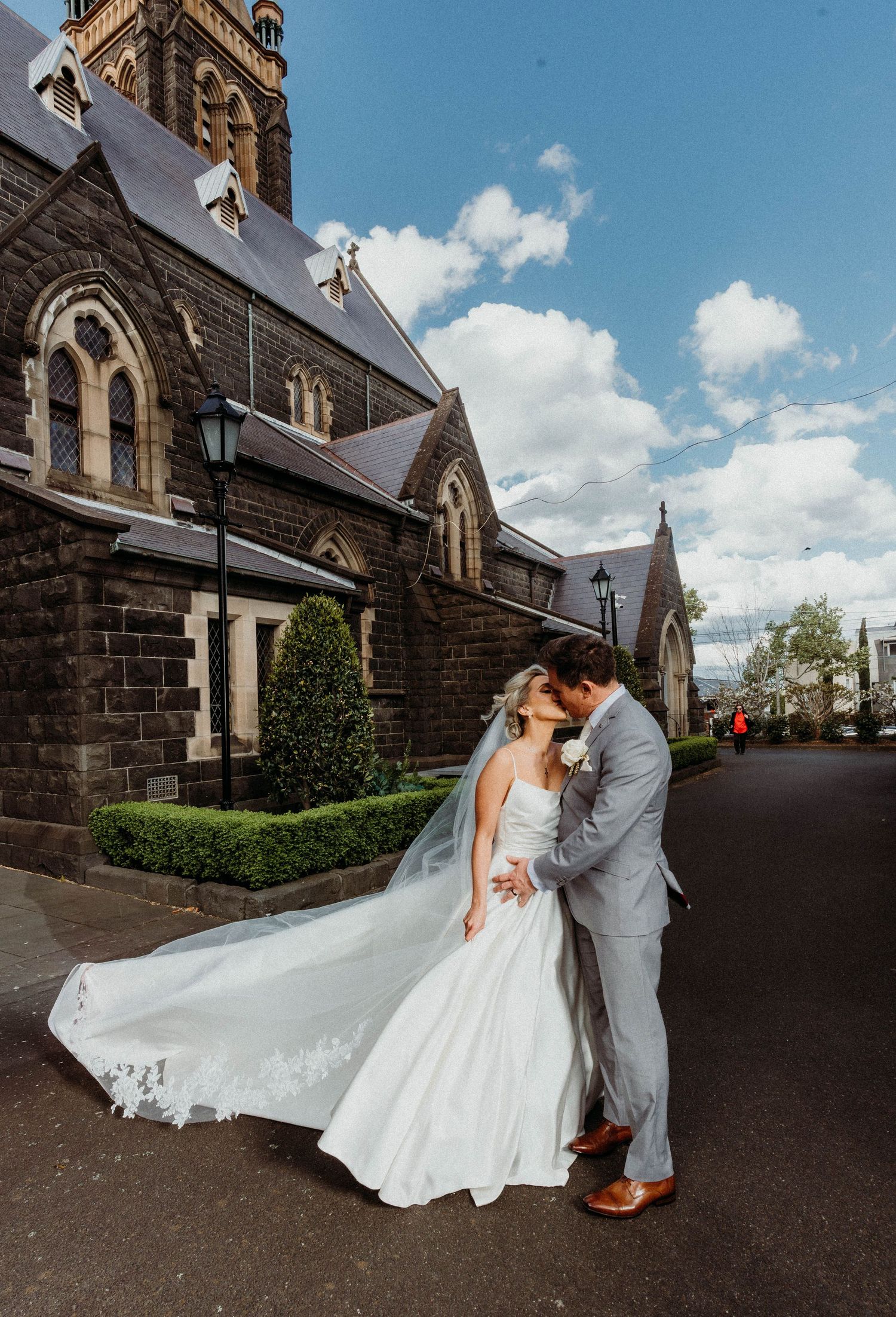 A romantic wedding scene outside a gothic stone church with arched windows on a sunny day with fluffy white clouds.