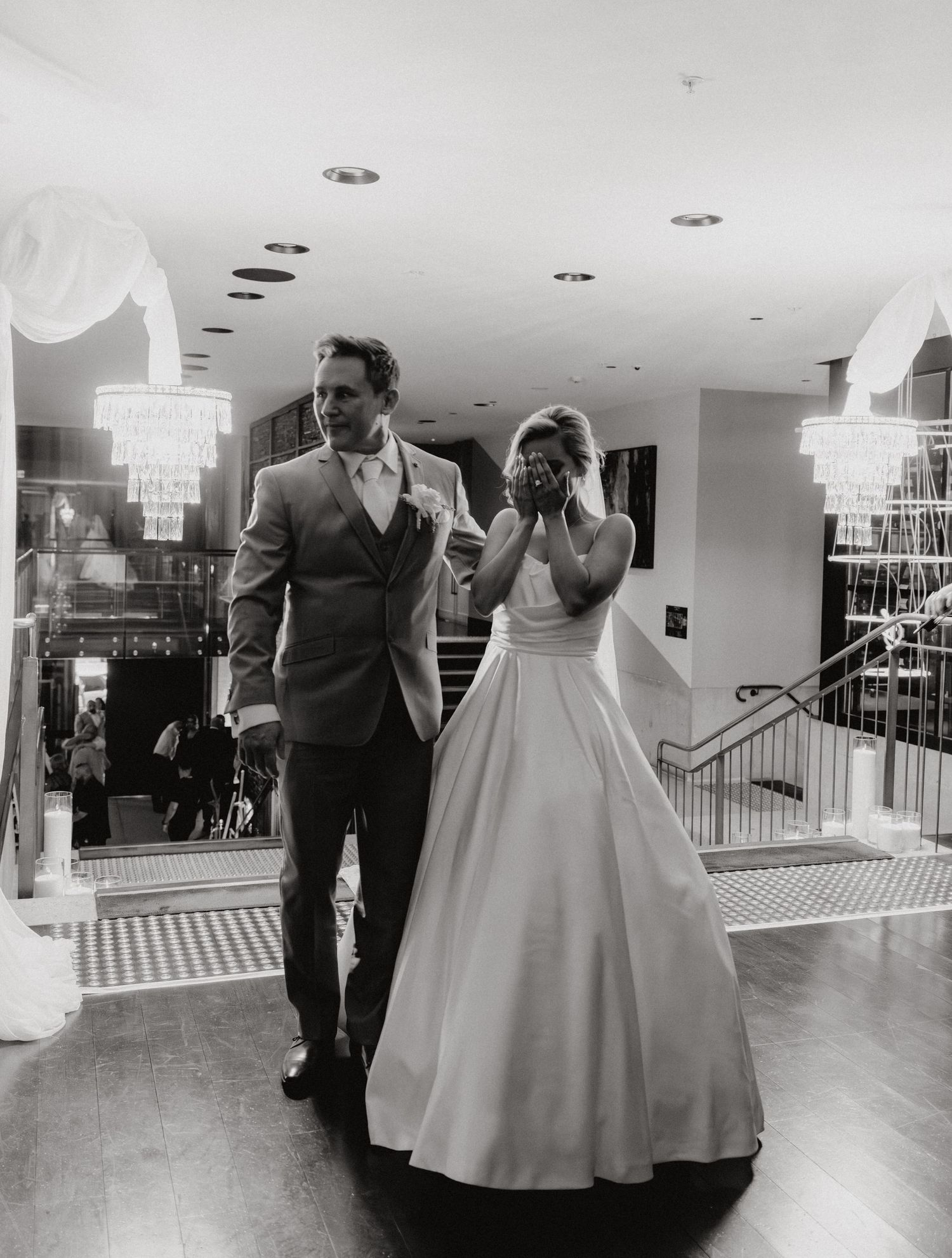Couple shares a romantic first dance moment in an elegant white ballroom with crystal chandeliers overhead.