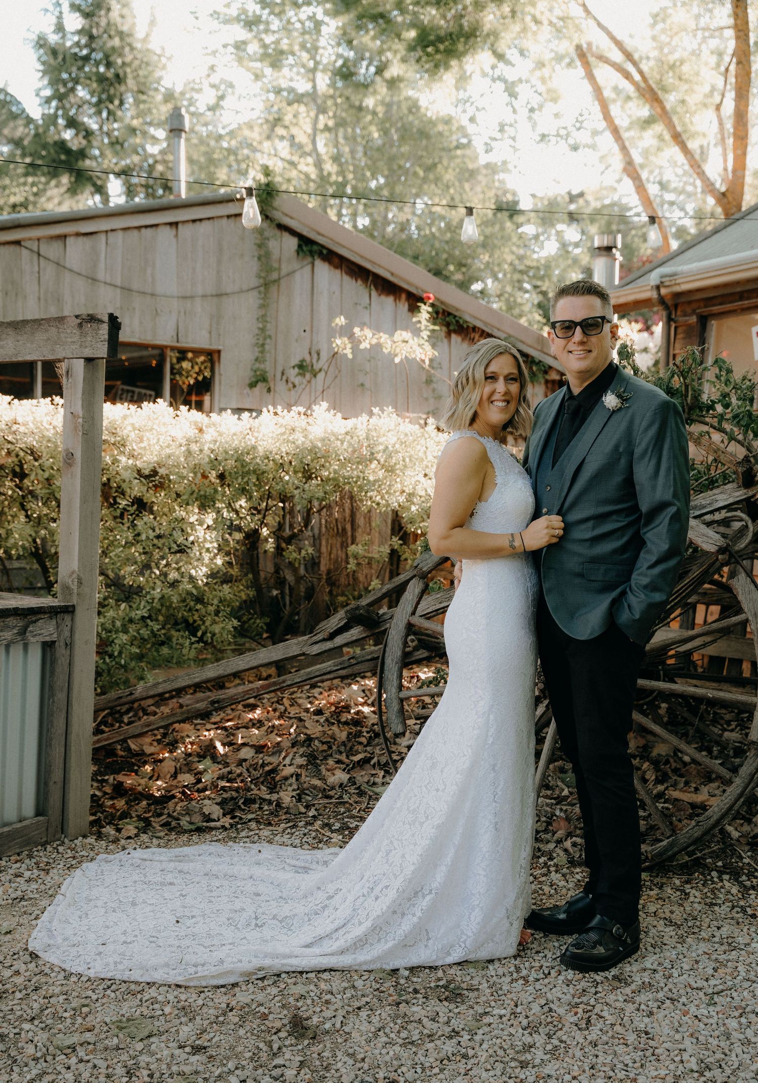 A couple poses together outdoors in formal attire against a rustic wooden fence at sunset.