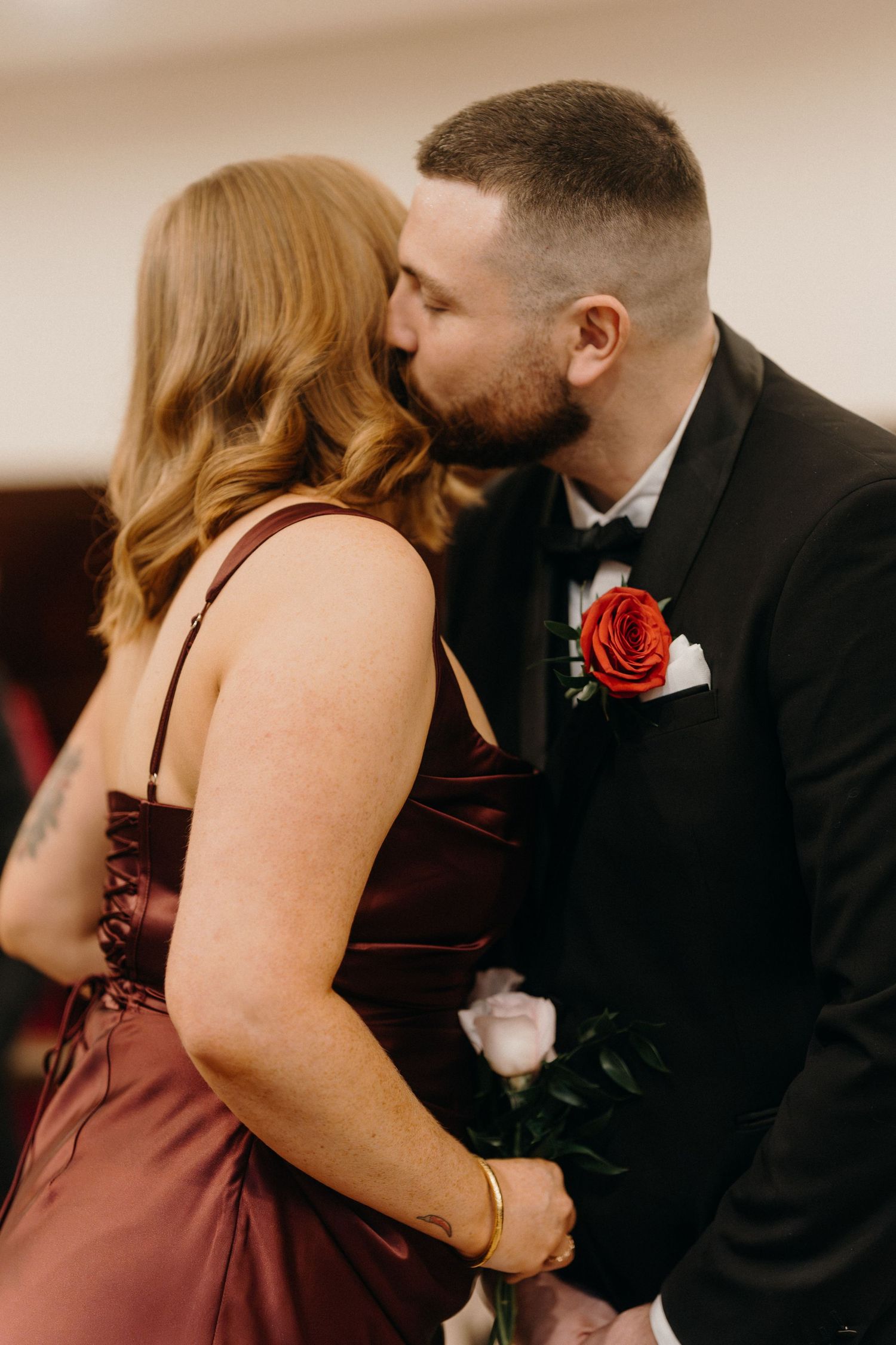 Couple sharing a romantic kiss at a wedding, groom in black tuxedo with red rose boutonniere, bride in burgundy satin gown.
