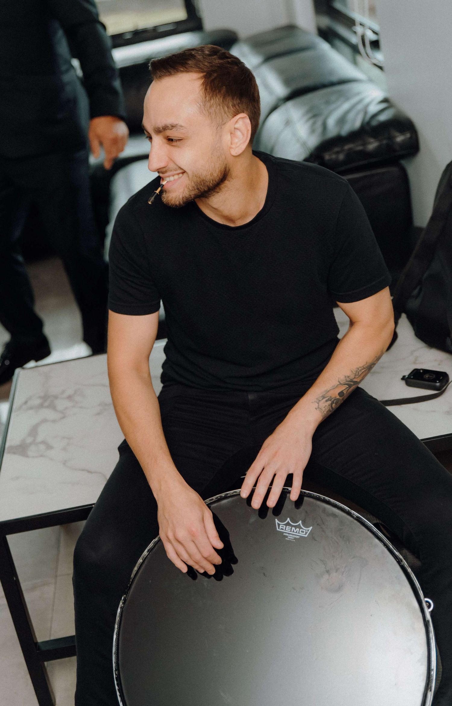 Person in black t-shirt smiling while sitting with drumming equipment at indoor studio.