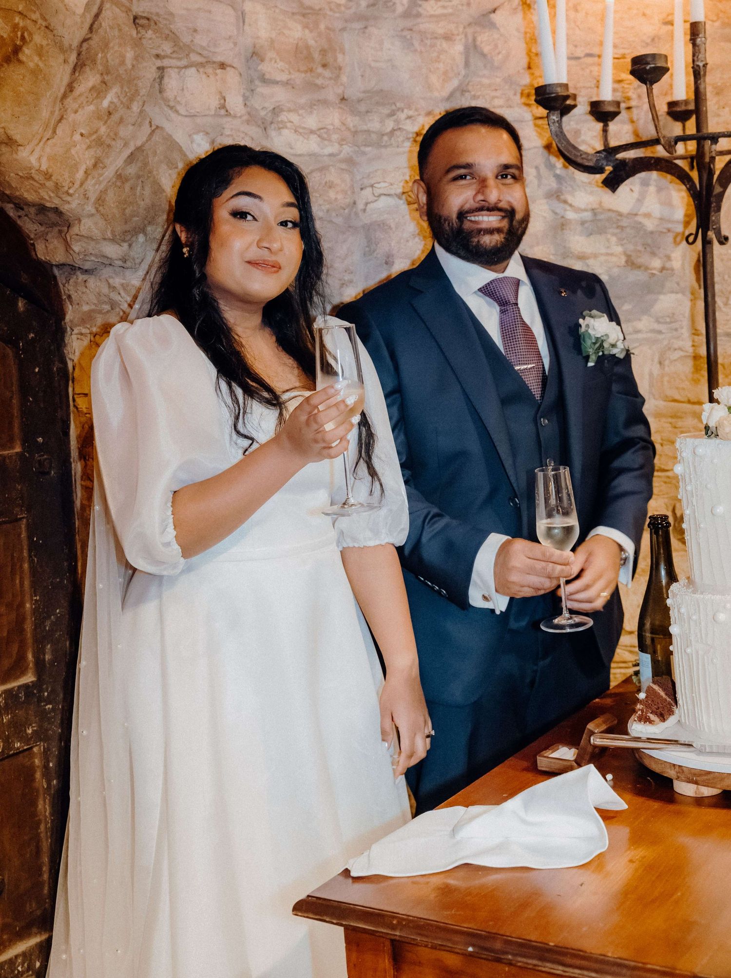 A couple in wedding attire holds champagne glasses and smiles during a wedding celebration in a rustic stone venue.