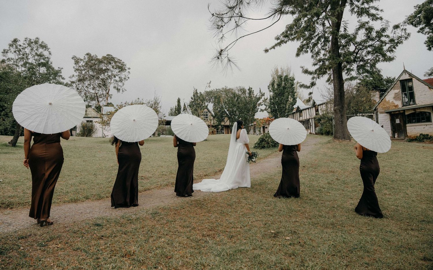 Creative wedding party photo with five people holding white umbrellas standing in a line on grassy field.