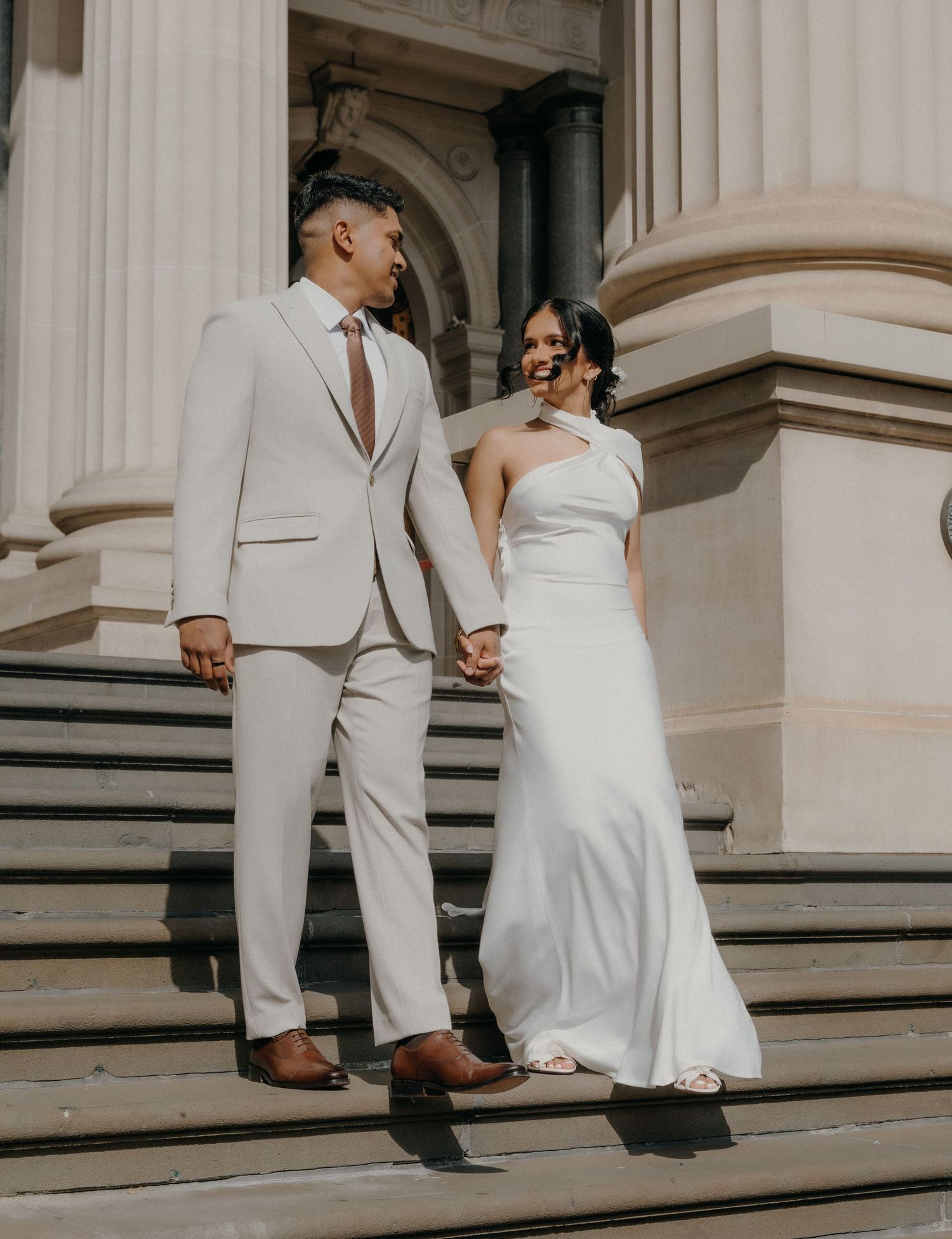 A couple in white wedding attire holds hands while walking up grand stone steps with elegant columns in the background.