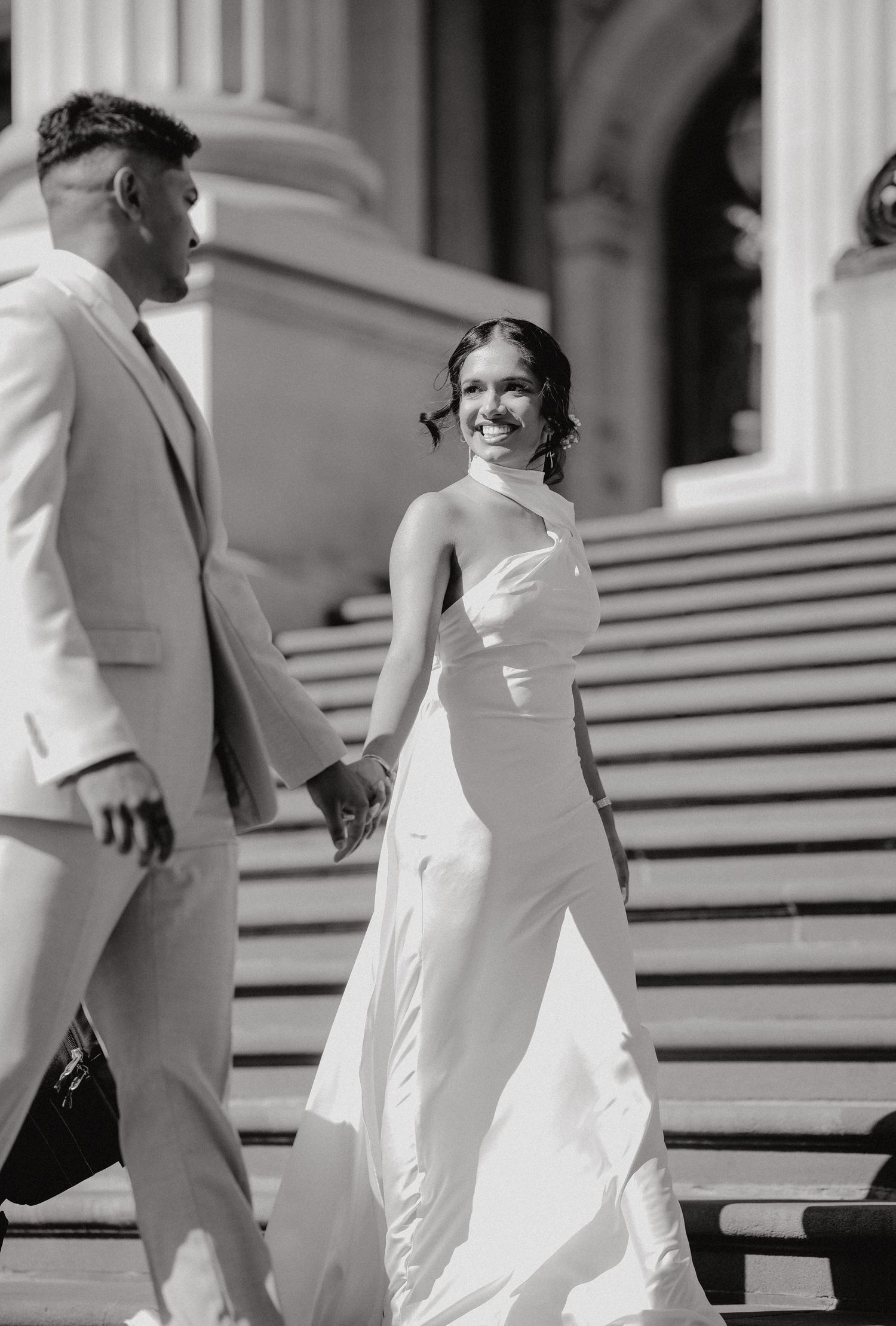 Elegant couple in white attire walking down grand stone steps during a formal occasion or wedding photoshoot.