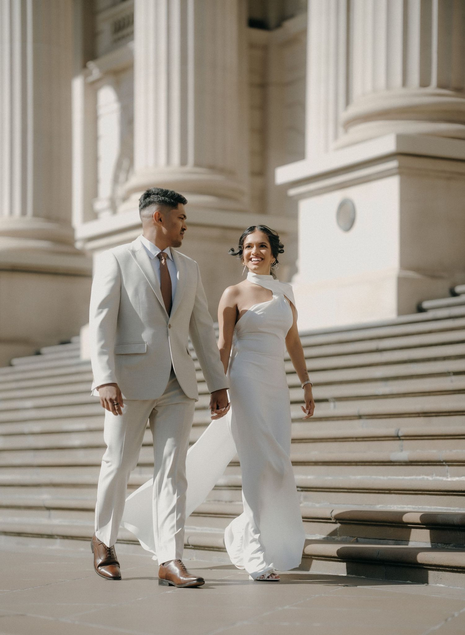 A couple in elegant white attire walks up grand marble steps in a classic architectural setting.
