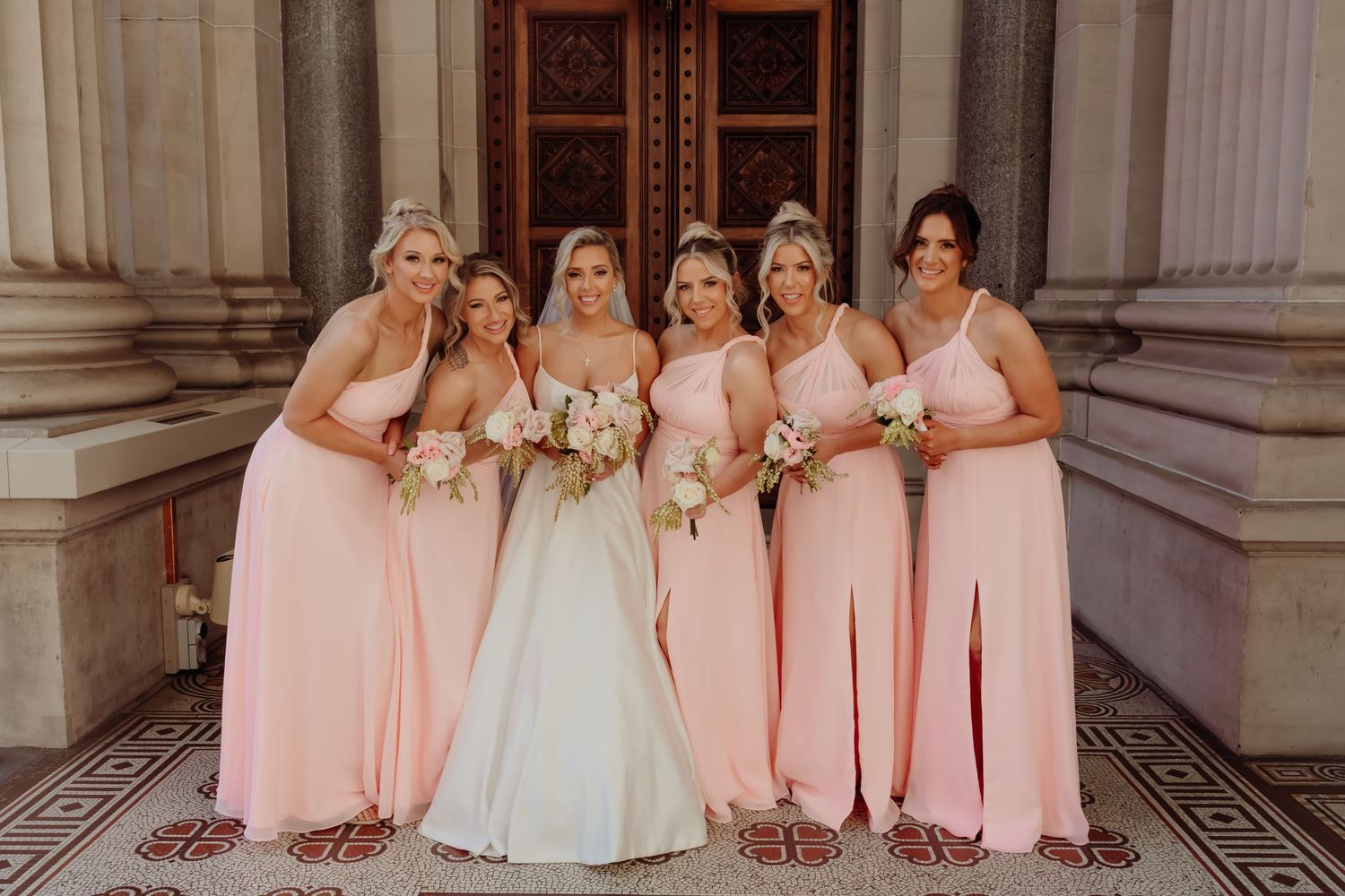 Wedding party in matching pink bridesmaid dresses poses together in an ornate hallway with marble columns.