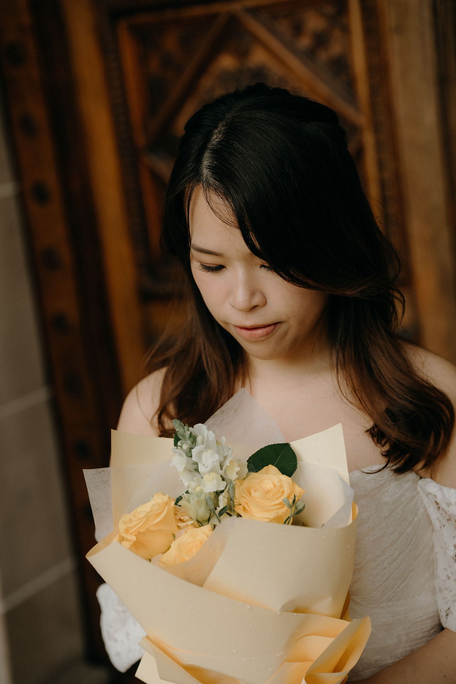 A person in a white dress holds a delicate bouquet of peach roses wrapped in cream-colored paper.