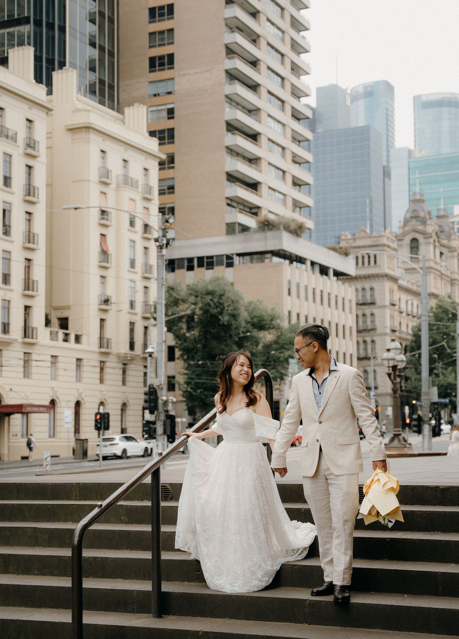 A couple in wedding attire descends stone steps in front of classic urban architecture and modern high-rise buildings.
