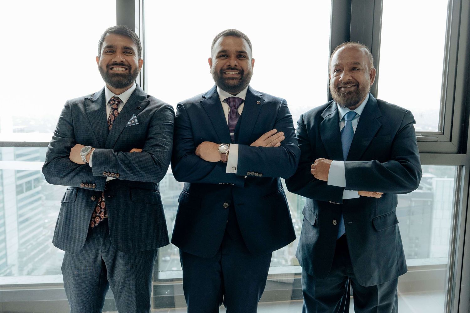 Three business professionals in dark suits stand with crossed arms, smiling confidently in bright office setting.