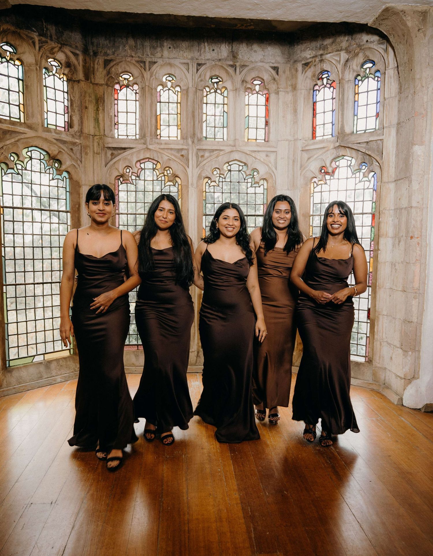 Five women in elegant black and brown evening gowns pose together in a stone room with tall arched windows.