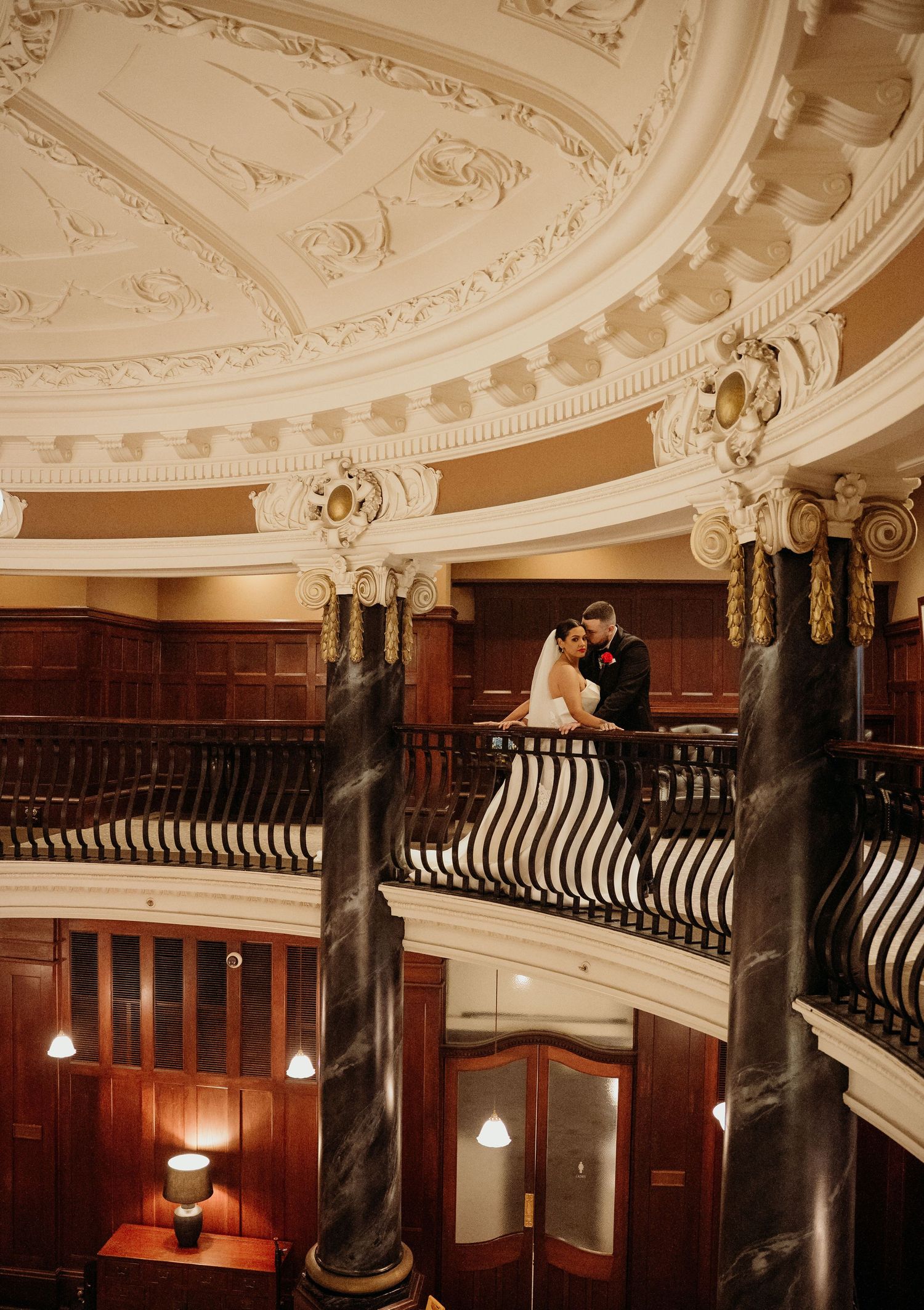 Bride and groom share a romantic moment on an ornate balcony with marble columns and decorative plasterwork ceiling.