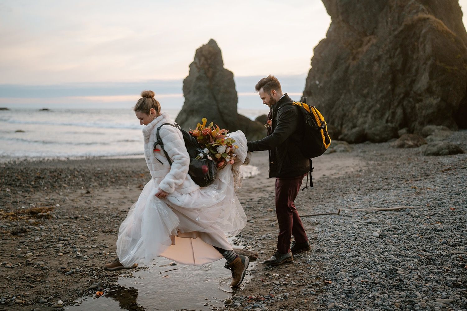 A couple walks along a rocky beach at sunset with the bride's white dress flowing in the ocean breeze.