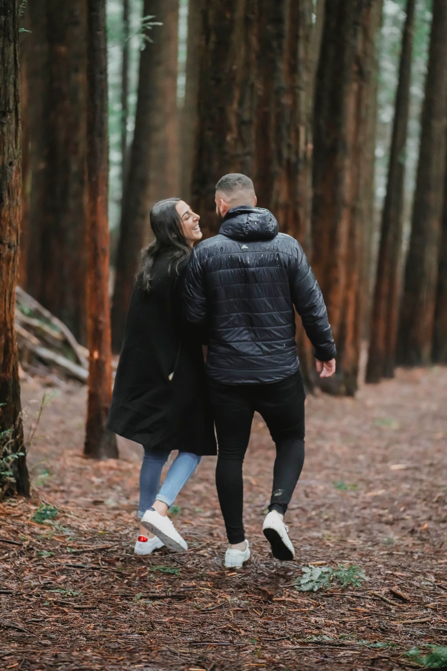 Two people in winter coats bend down to examine something on a forest trail covered in fallen pine needles.