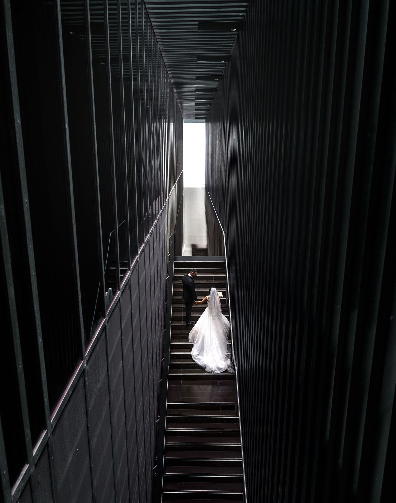 A dramatic view down a dark industrial hallway with bright light at the end, featuring vertical metal bars along the walls.