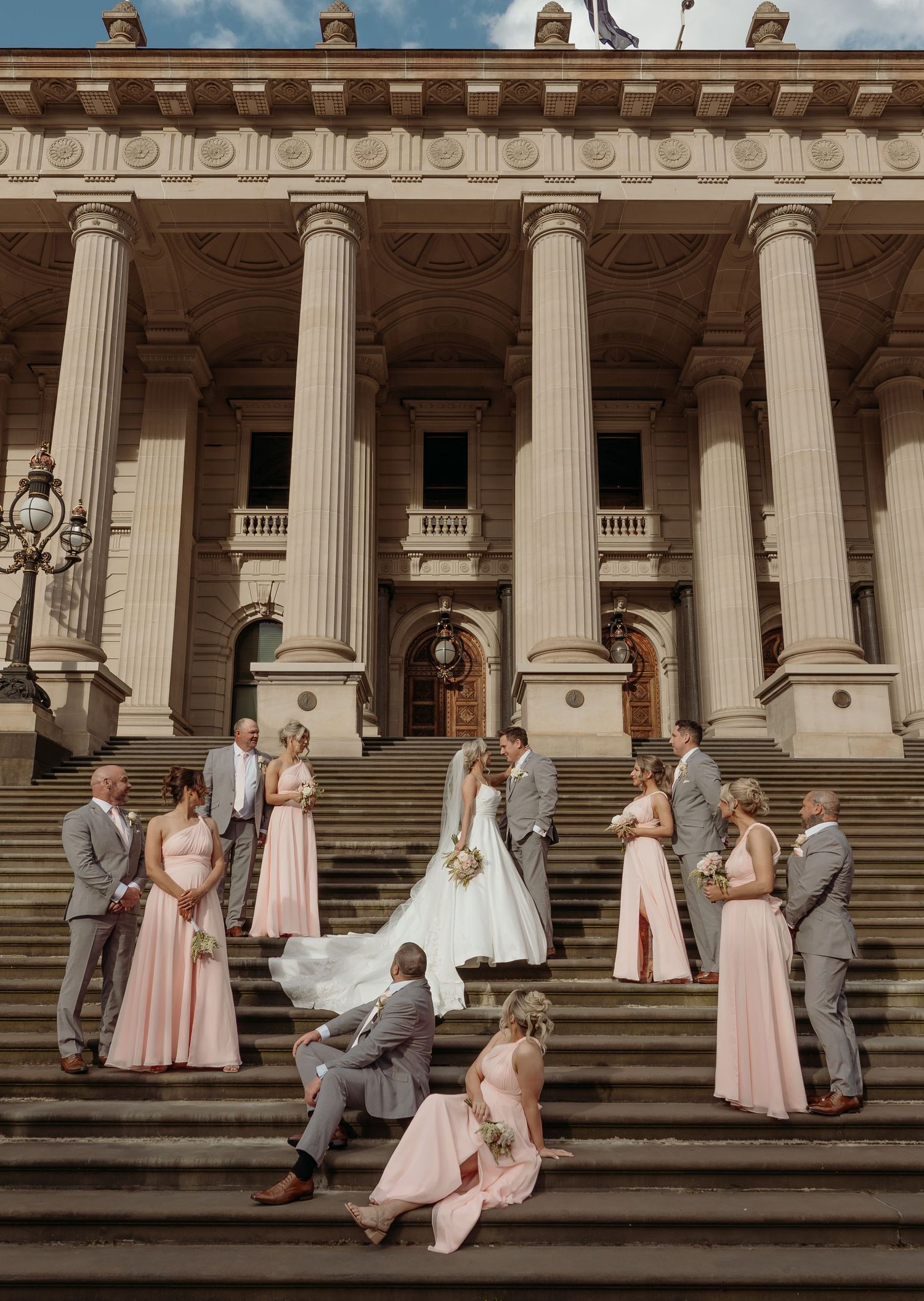 Wedding party poses on grand stone steps with white columns of historic building in elegant pink and gray attire.