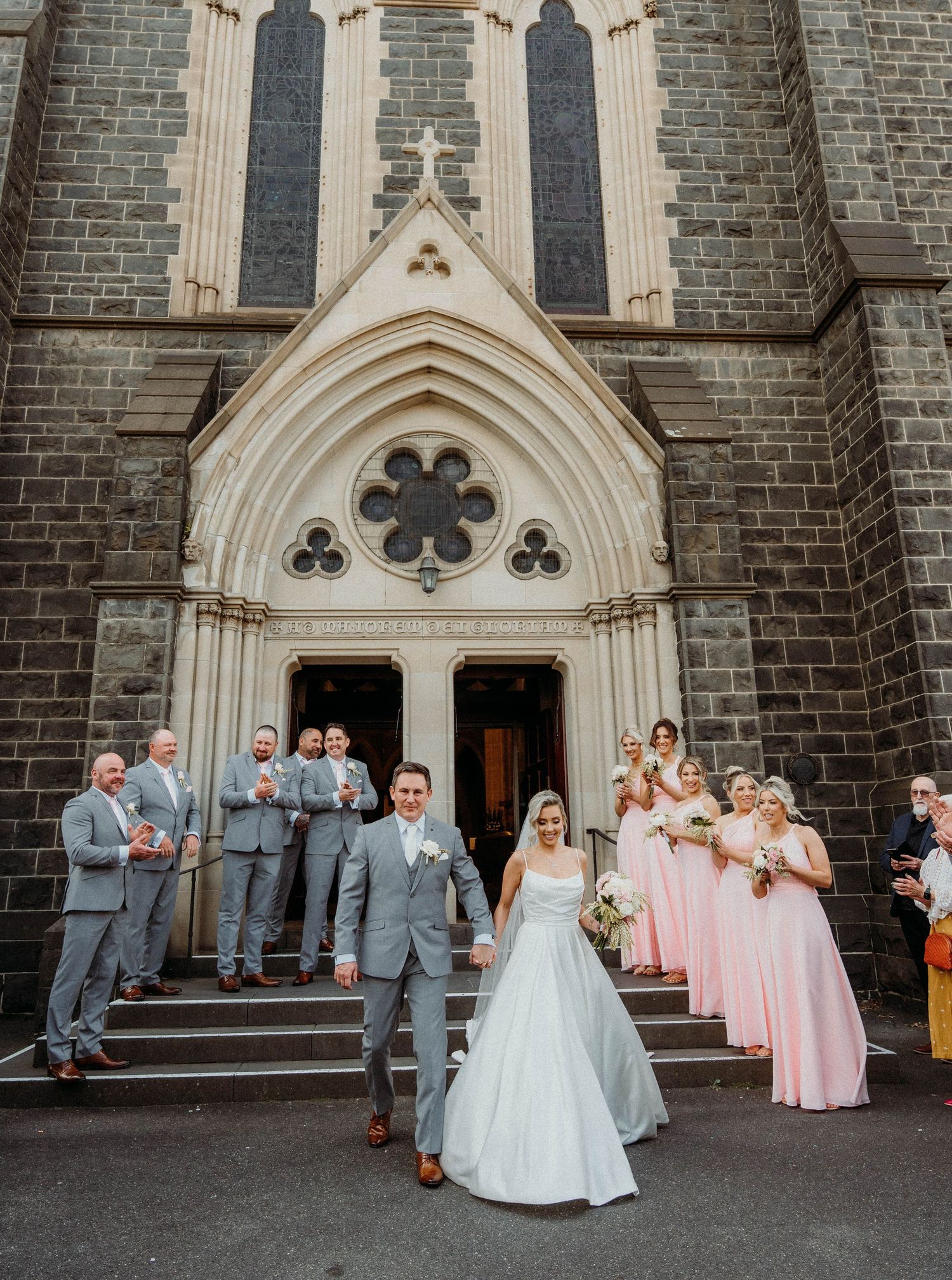 Wedding party in pink dresses and gray suits gather outside a gothic stone church with ornate architecture.