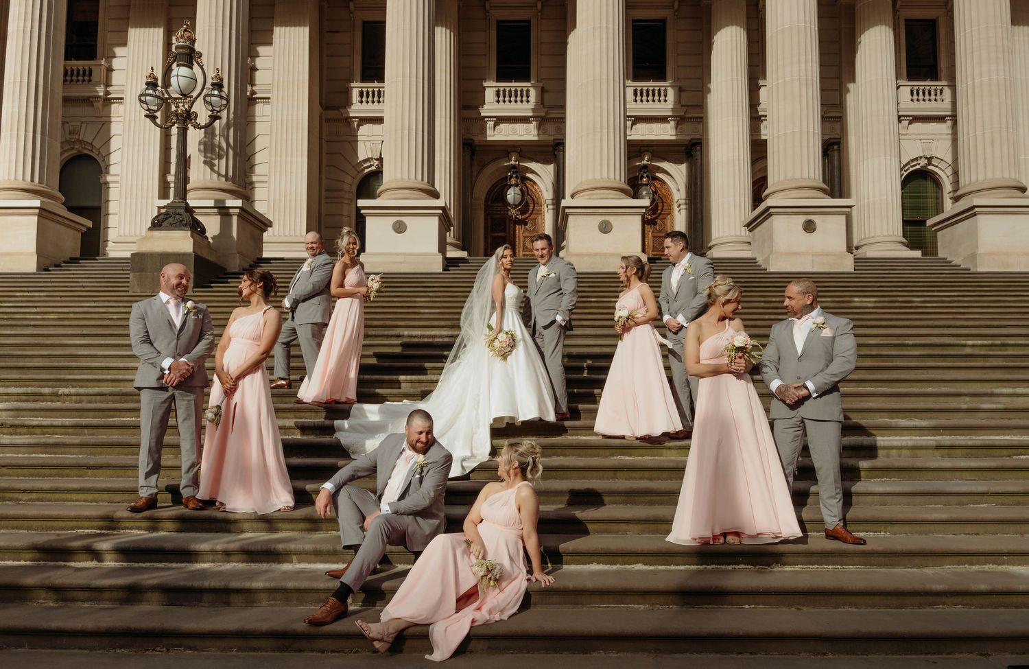 Wedding party members pose on grand stone steps with white columns of historic building in background.