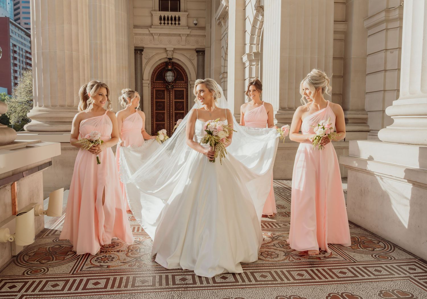 A wedding party in blush pink bridesmaid dresses poses on elegant stone steps of a historic building.