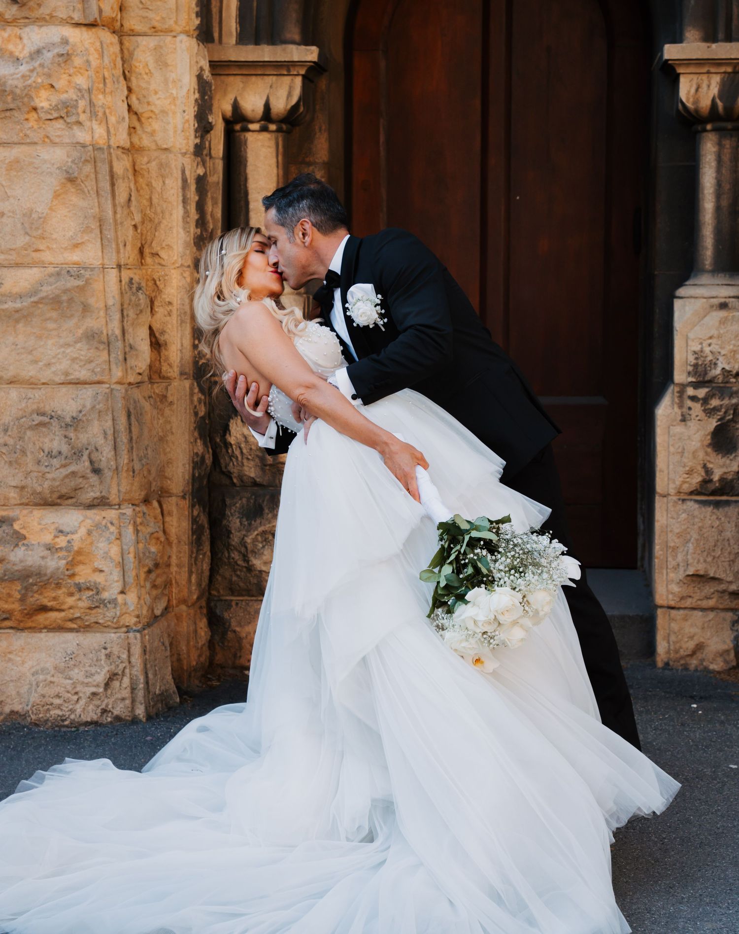 A romantic wedding couple shares an intimate kiss against a rustic stone archway backdrop.