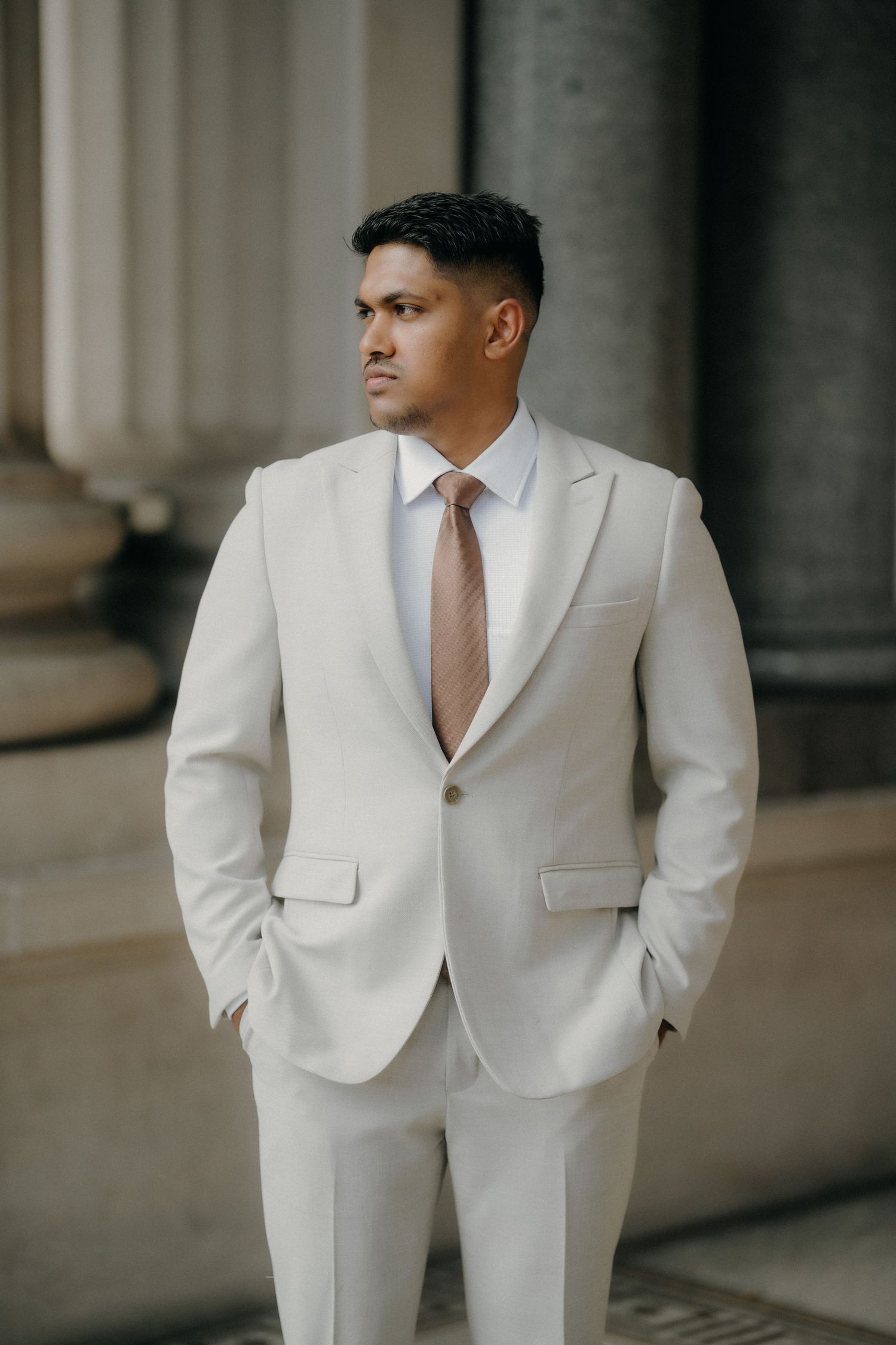 Professional portrait of someone wearing a crisp white suit with a pink tie standing against classical stone columns.