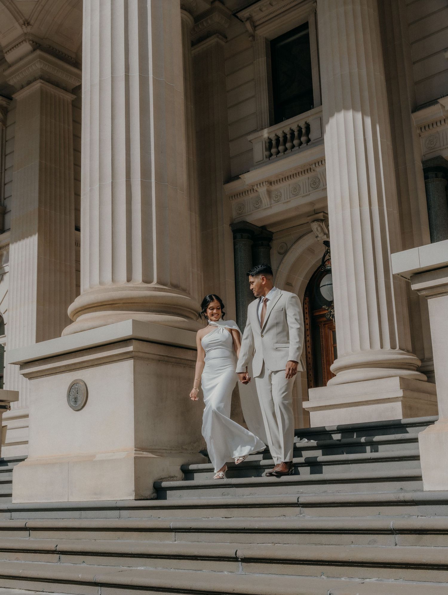 A couple poses on the steps of a grand building with white columns in an elegant wedding photo shoot.