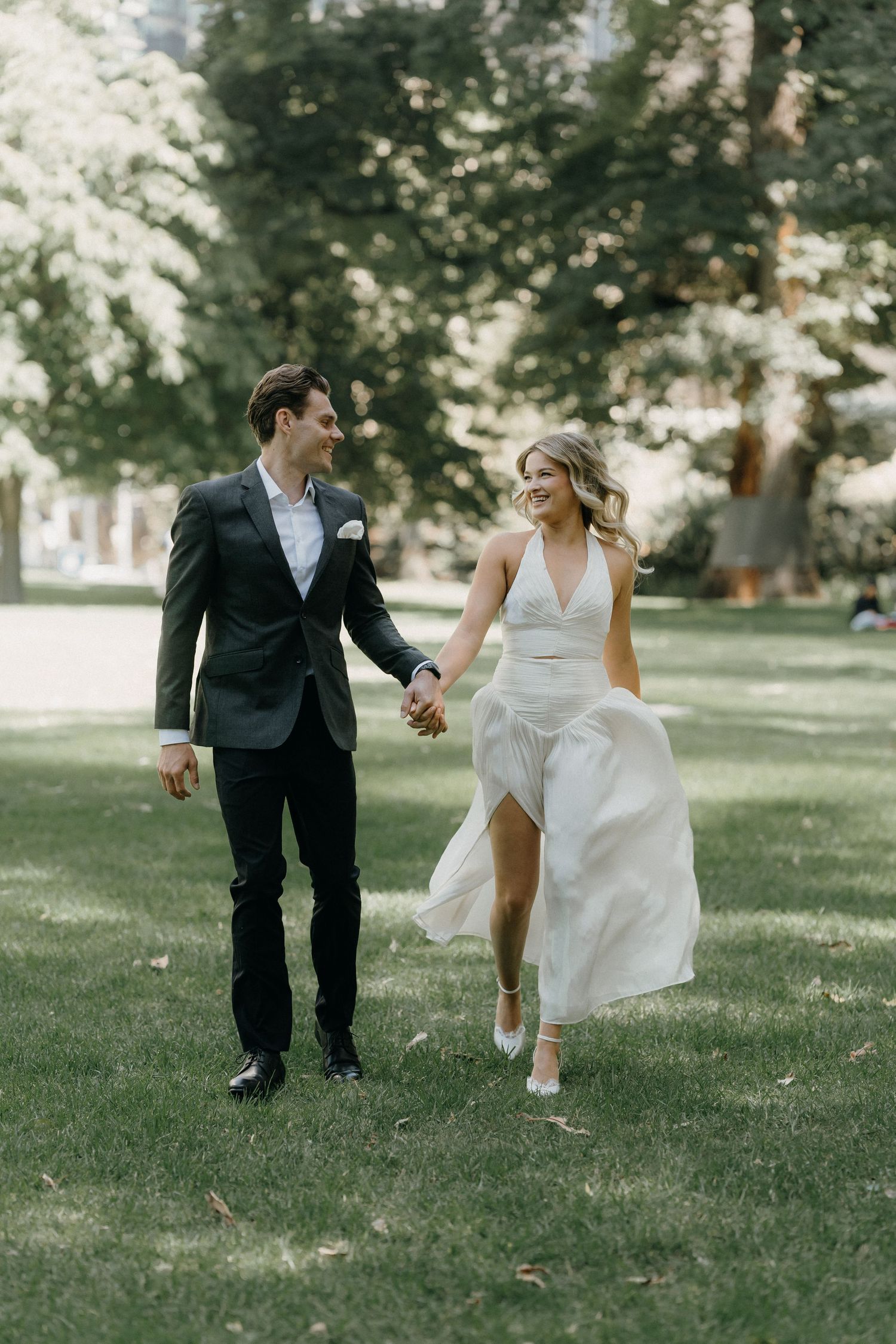 A smiling bride and groom walk hand-in-hand across a lush green lawn surrounded by trees on their wedding day.