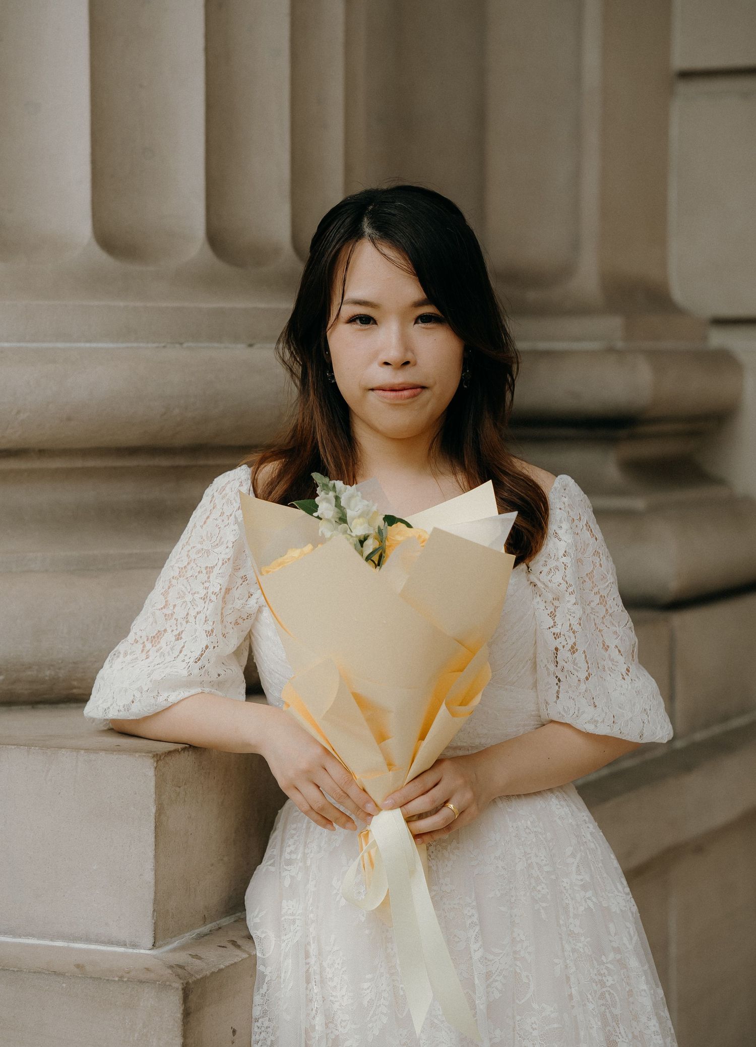 A person in a white lace dress poses elegantly against classical stone columns in a romantic bridal portrait.