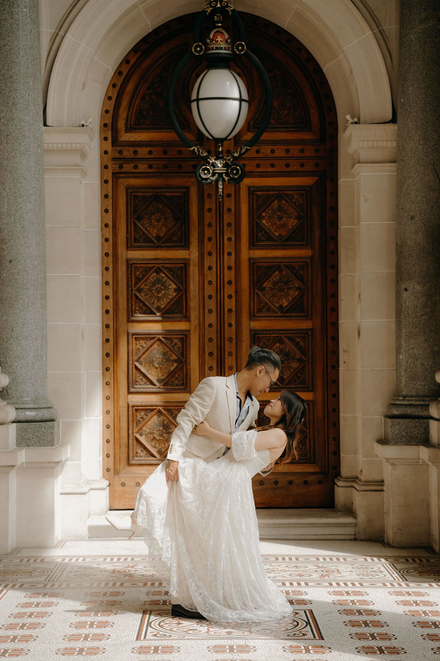 Romantic couple in wedding attire share intimate moment in front of ornate wooden doors at historic building entrance.