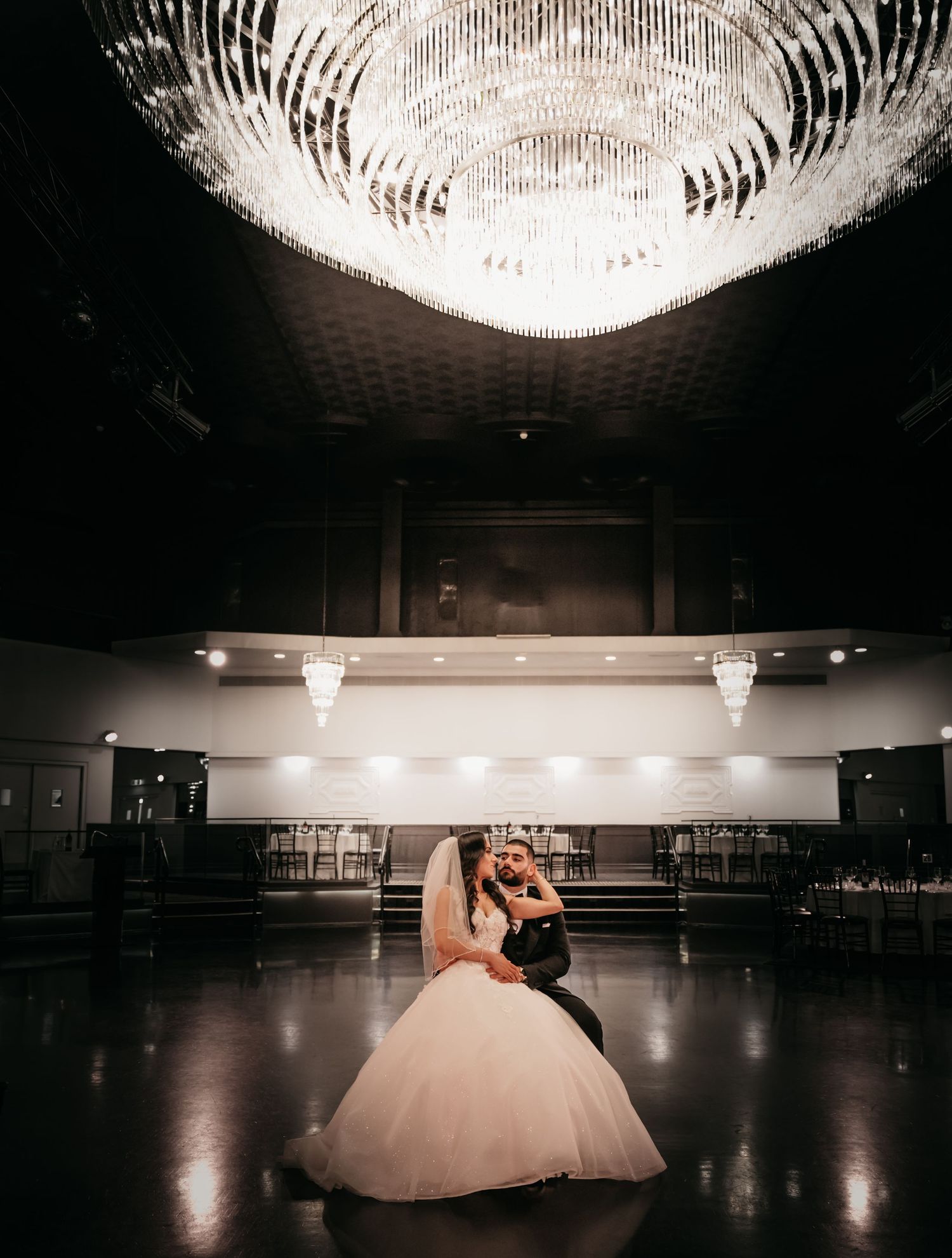 A couple dances beneath an elegant crystal chandelier in a grand ballroom with polished floors and dim lighting.