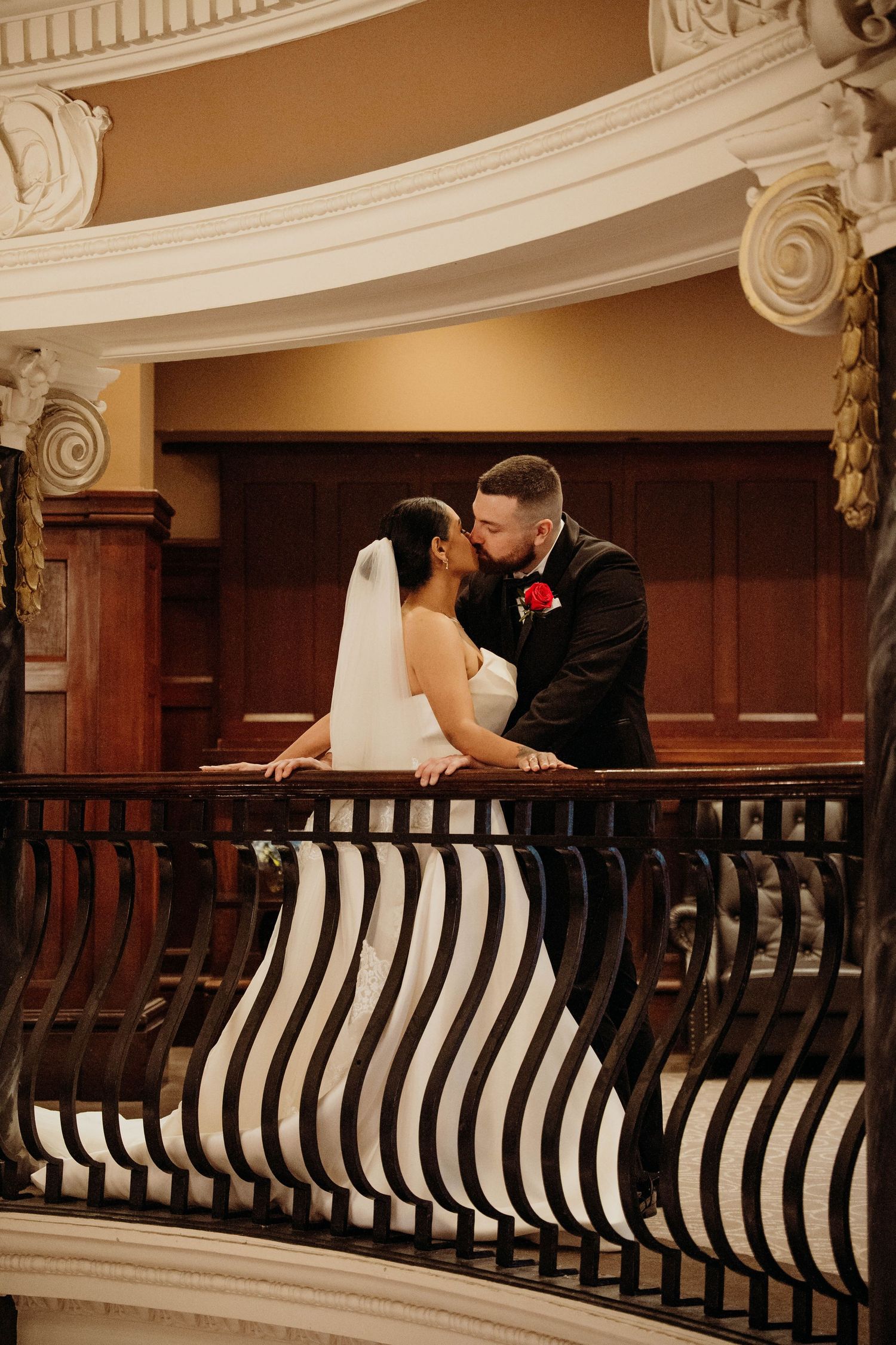 Bride and groom share a romantic kiss on an elegant balcony with striped railings in a grand venue.