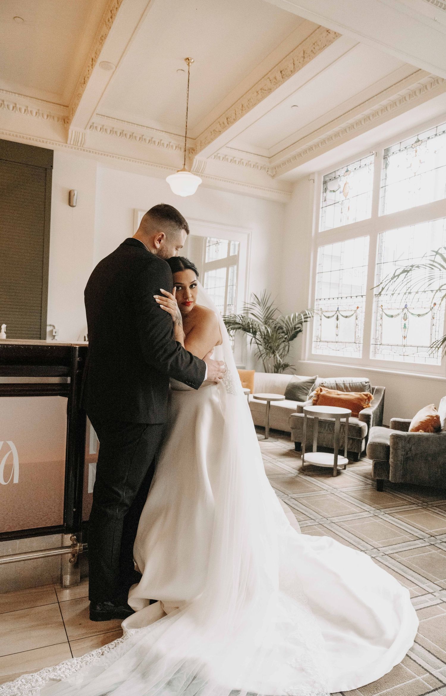 A bride and groom share a romantic embrace in an elegant sunlit room, her white gown trailing gracefully on the floor.