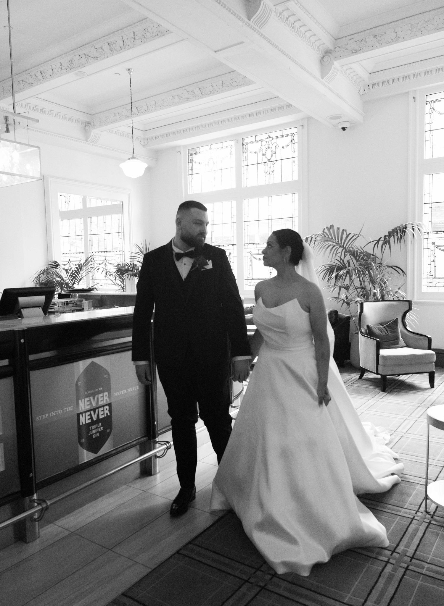 Black and white photo of a bride and groom holding hands in an elegant venue with ornate ceilings.