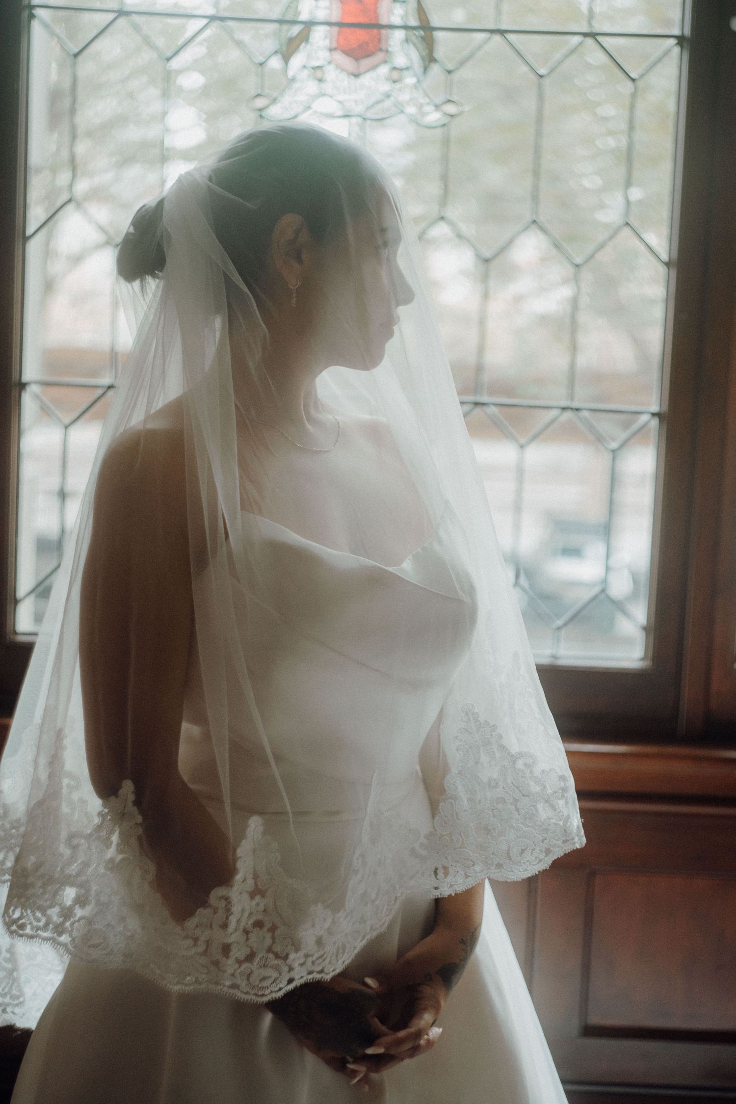 Bride in white lace-trimmed veil and strapless gown stands by stained glass window in soft natural light.