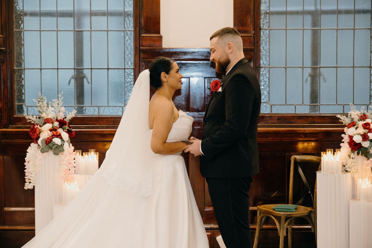 Bride and groom holding hands at wedding altar with red floral arrangements and candles in elegant venue.