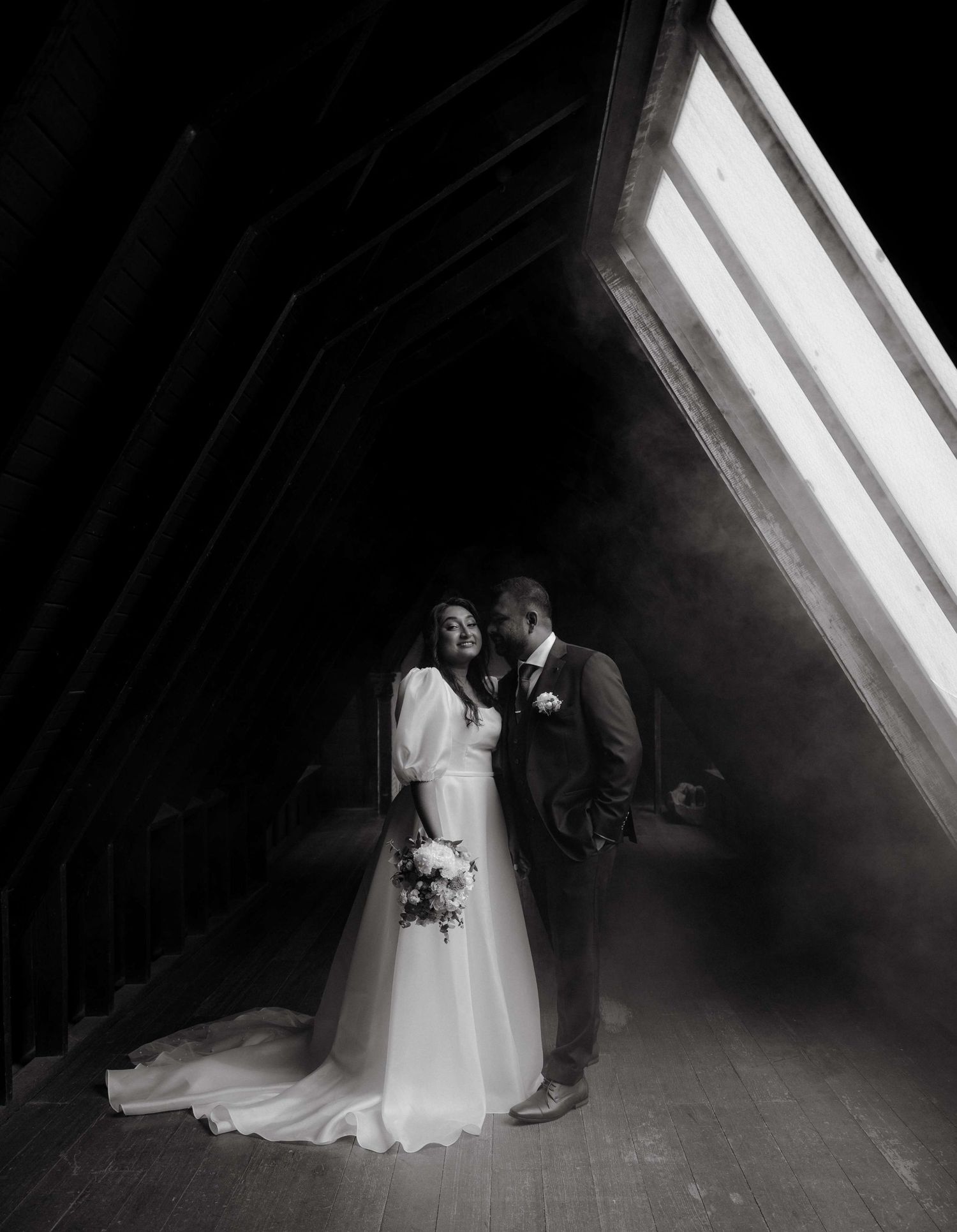 A dramatic black and white wedding photograph of a couple standing in an attic with natural light streaming through a window.