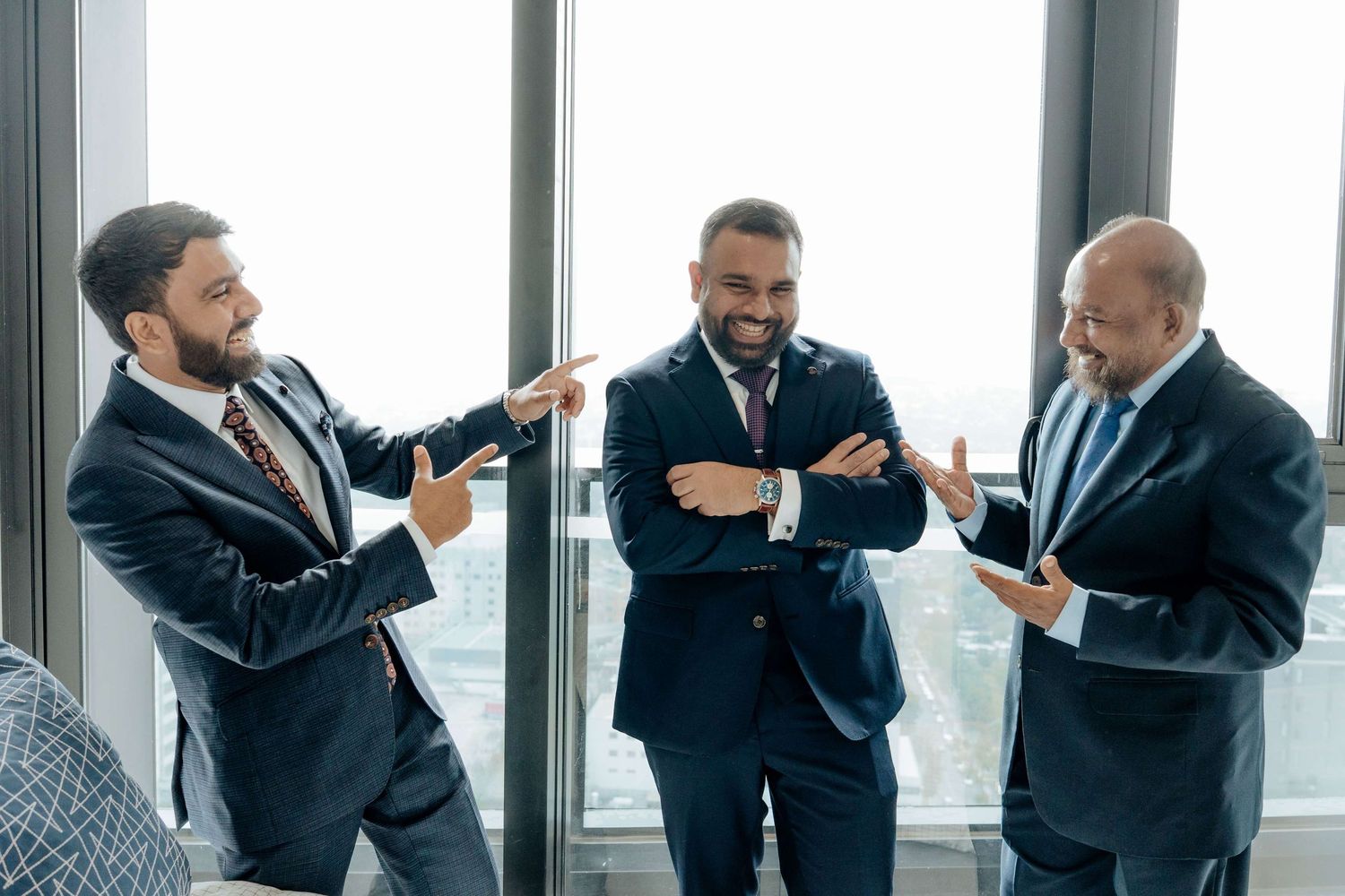 Business people in dark suits gesturing and interacting during an animated office conversation near large windows.