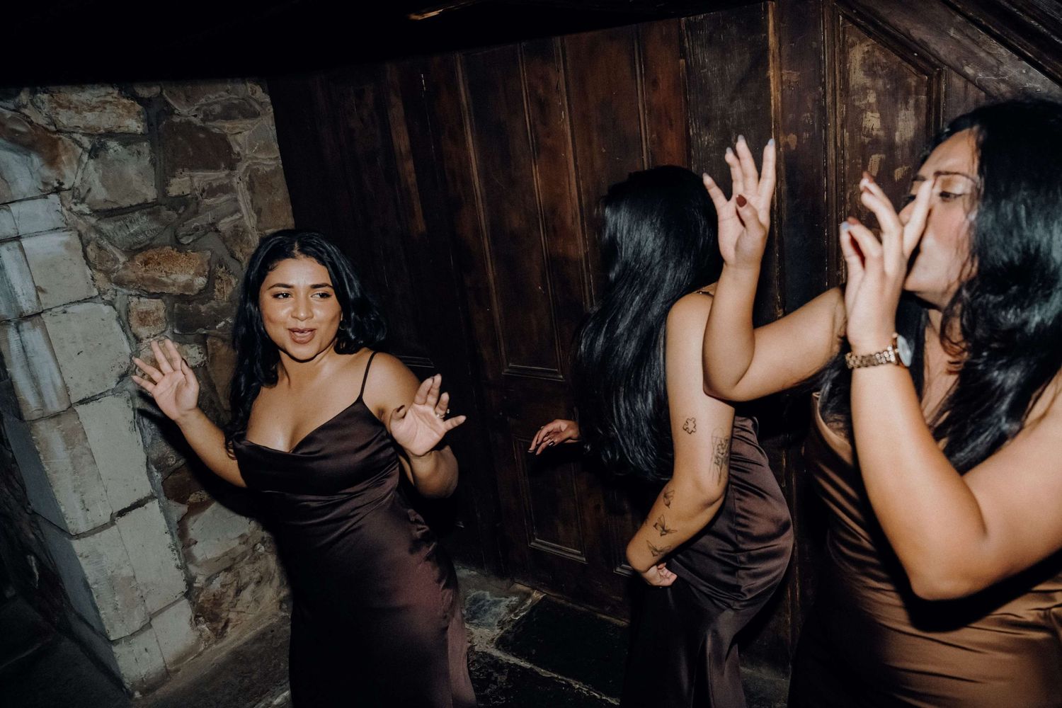 People in formal brown dresses dancing and celebrating at a nighttime party in a dimly lit rustic venue.