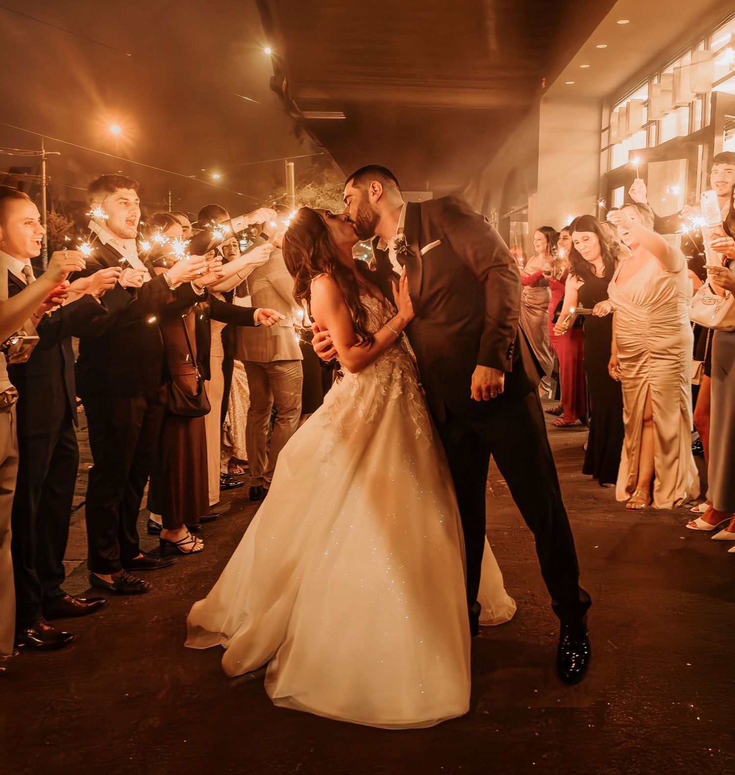 A couple share a romantic kiss during their sparkler exit as wedding guests line the path on a warm glowing evening.