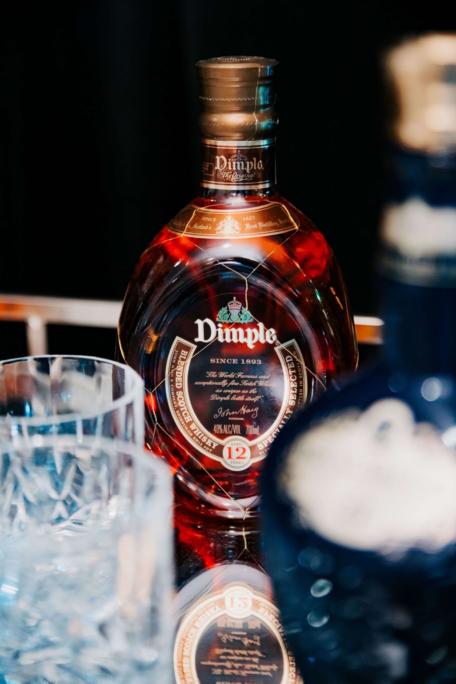 Crystal glasses and bottles of Dimple whisky on dark surface with dramatic lighting showcasing the iconic bottle design.