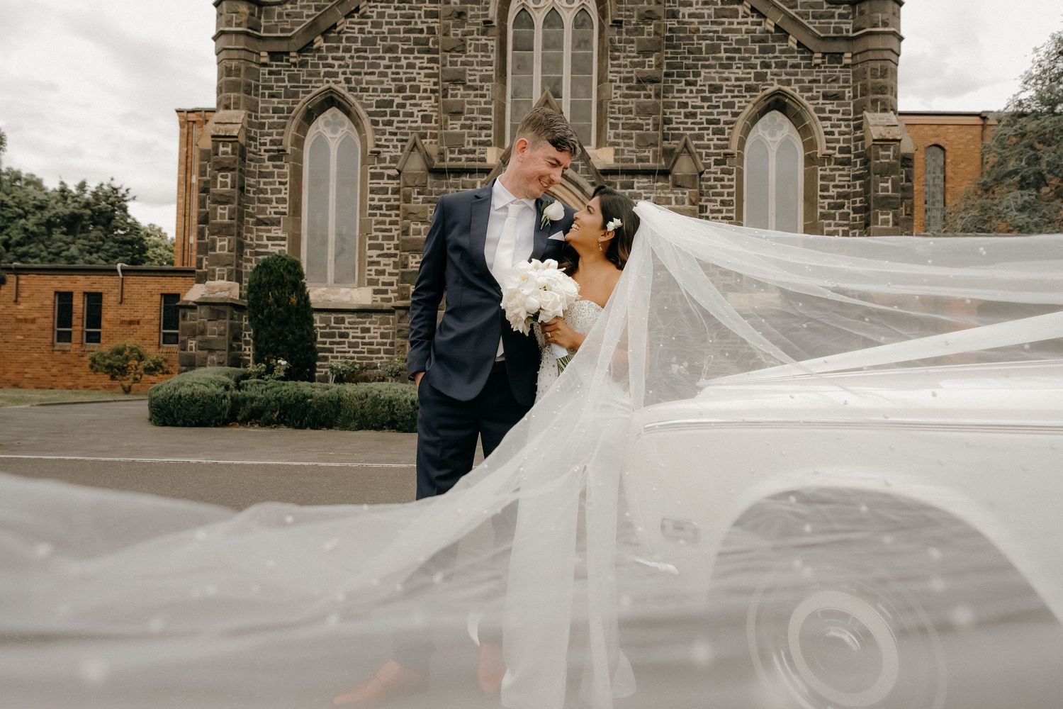 A flowing white wedding veil blows gracefully in the wind outside a stone church building.
