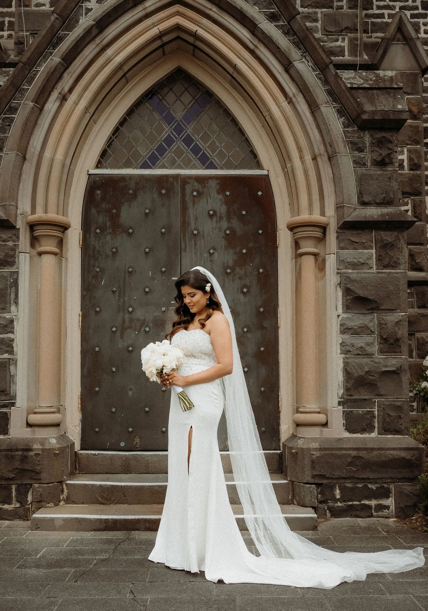 A bride in a flowing white wedding dress and veil poses gracefully against a gothic stone church entrance with arched doorways.