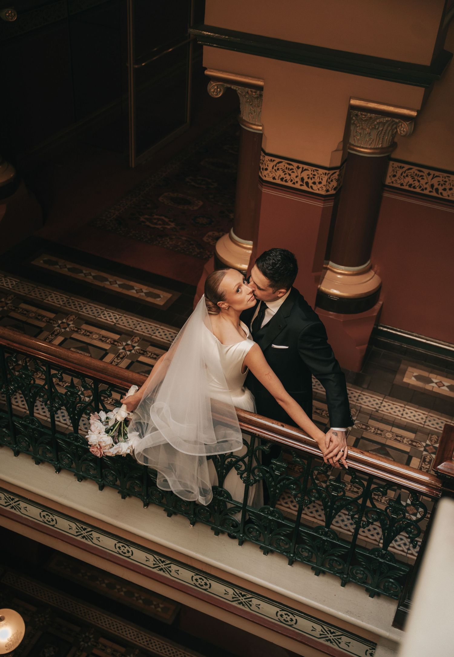 Bride and groom share a romantic kiss on an ornate balcony, captured from above, bride holding white bouquet.