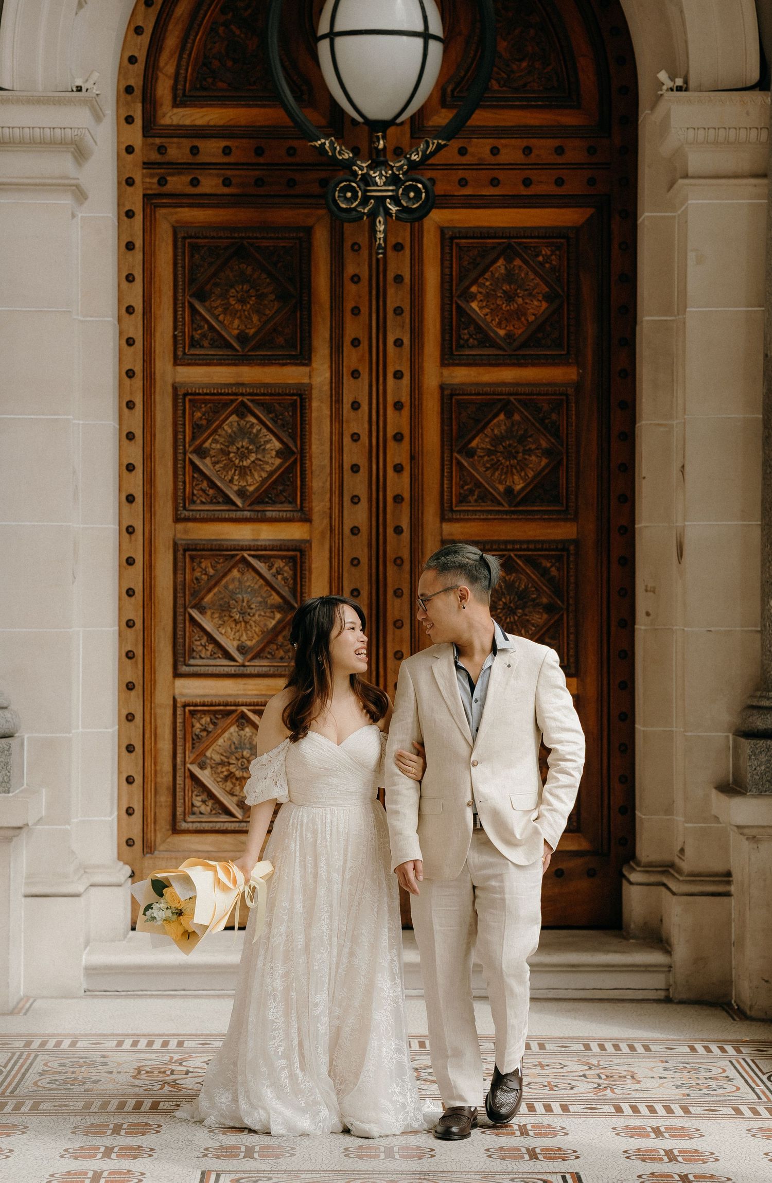 A romantic wedding couple poses in front of ornate wooden doors at a grand historic building.
