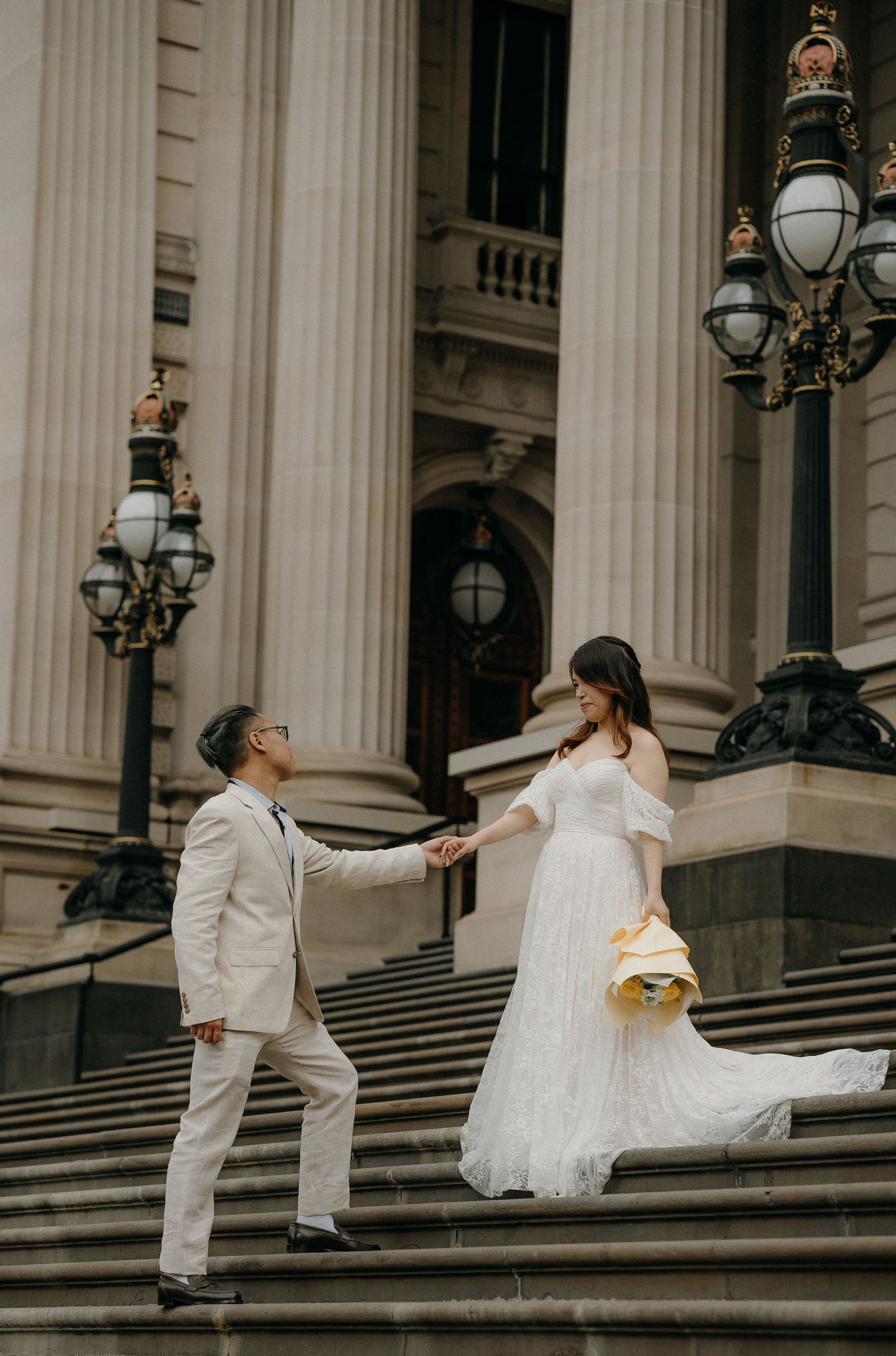A couple in white wedding attire dances gracefully on grand stone steps with ornate columns and lampposts in the background.