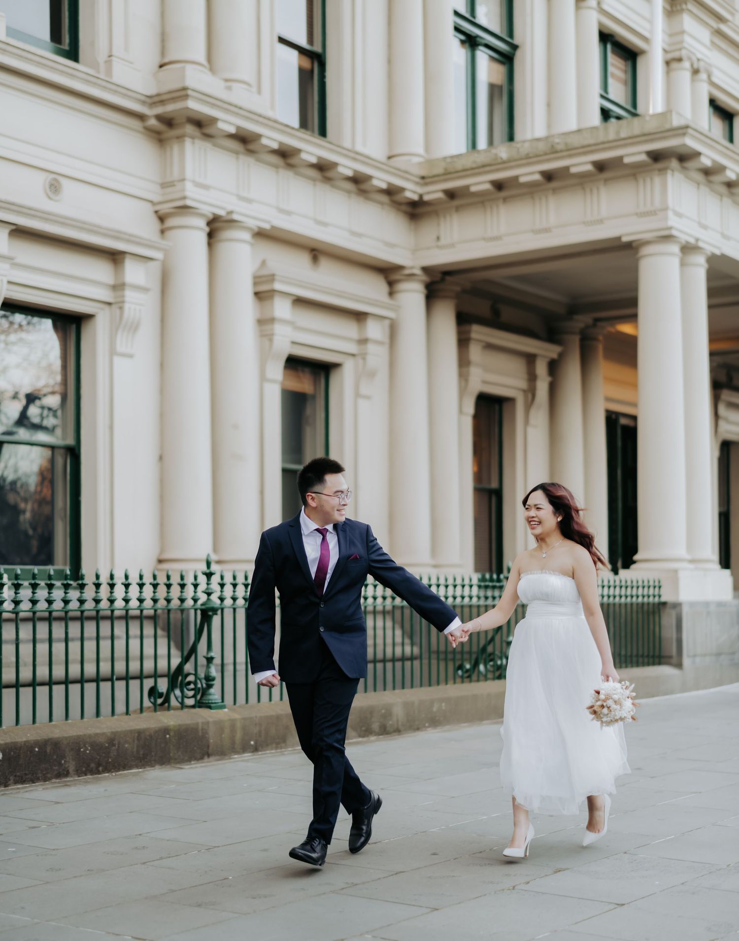 A couple in wedding attire walks together and holds hands outside a grand historic building with white columns.